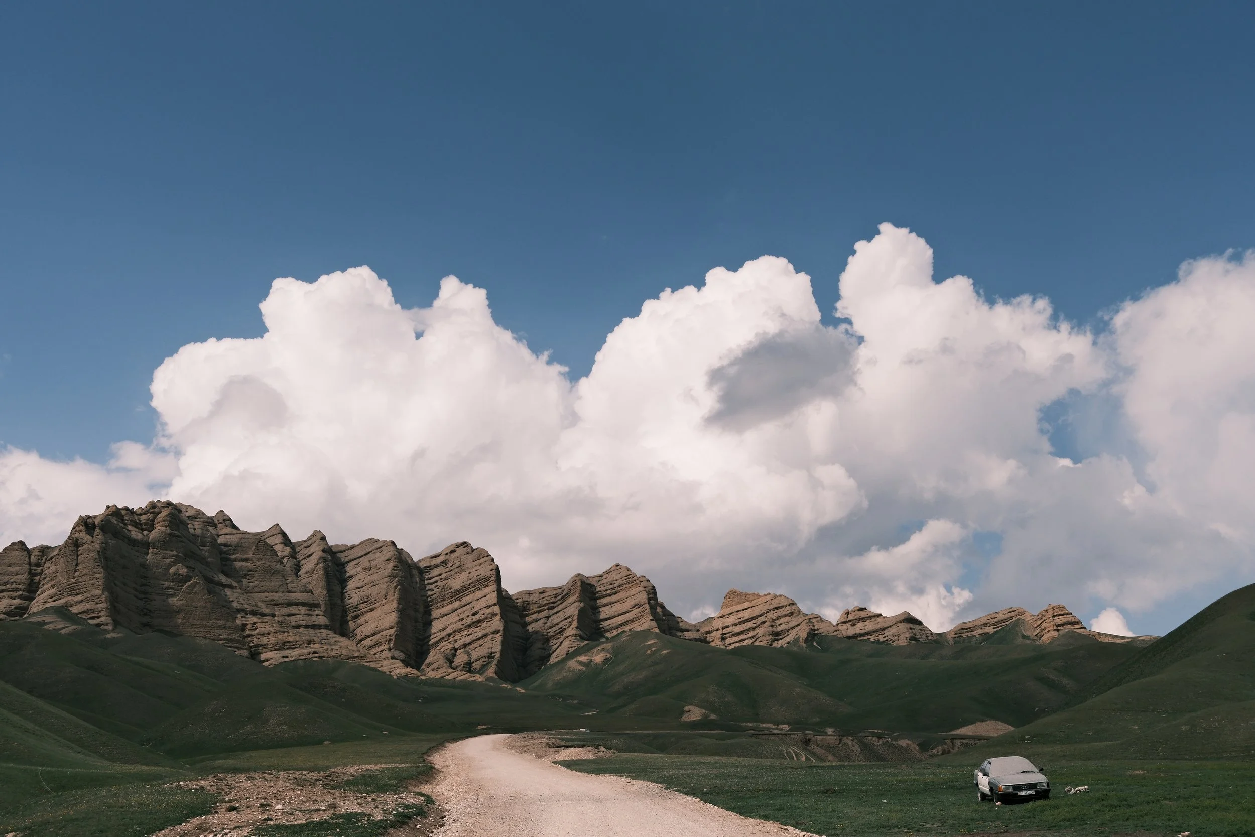 Landscape with mountains, a grassy valley, a winding road and a vehicle in the meadow, under a sky with large white clouds.
