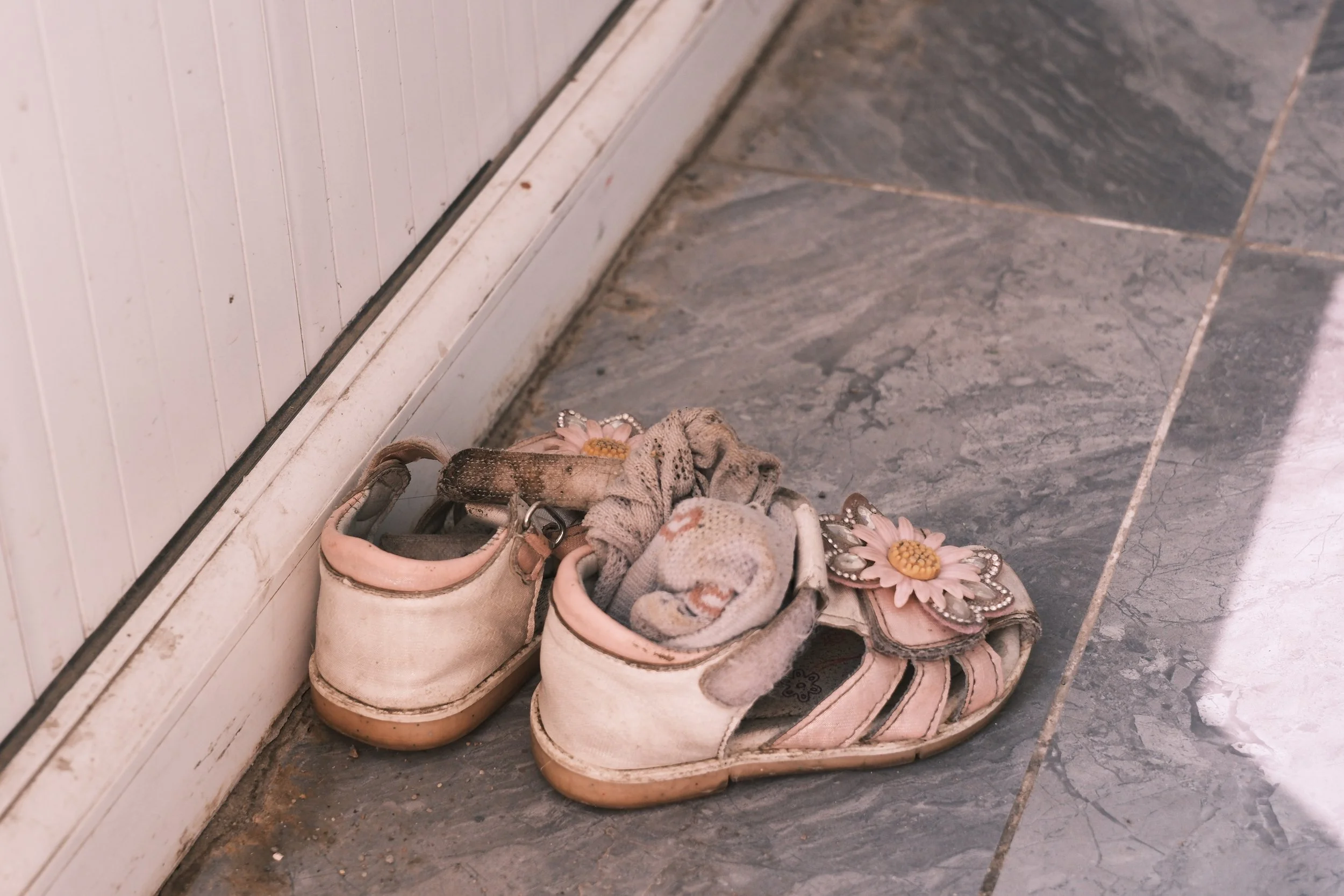 A pair of old, dirty children's shoes with flower decorations on the floor, next to a white wall and a white plinth.