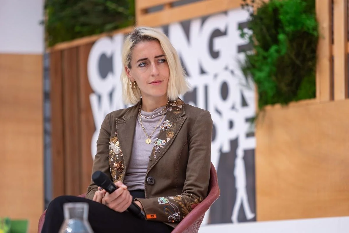 Young woman with blonde, shoulder-length hair sits on a chair and holds a microphone in her hand. In the background is a mural with black and white letters.