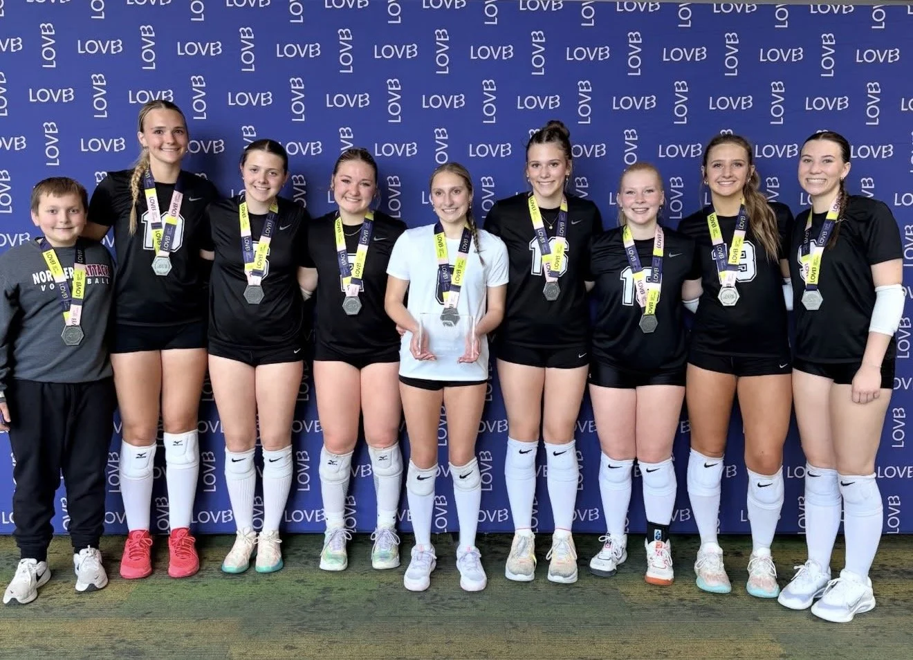 A girls' volleyball team celebrating with medals and a trophy at a victory event, standing in front of a blue backdrop with the word LOVE repeated.