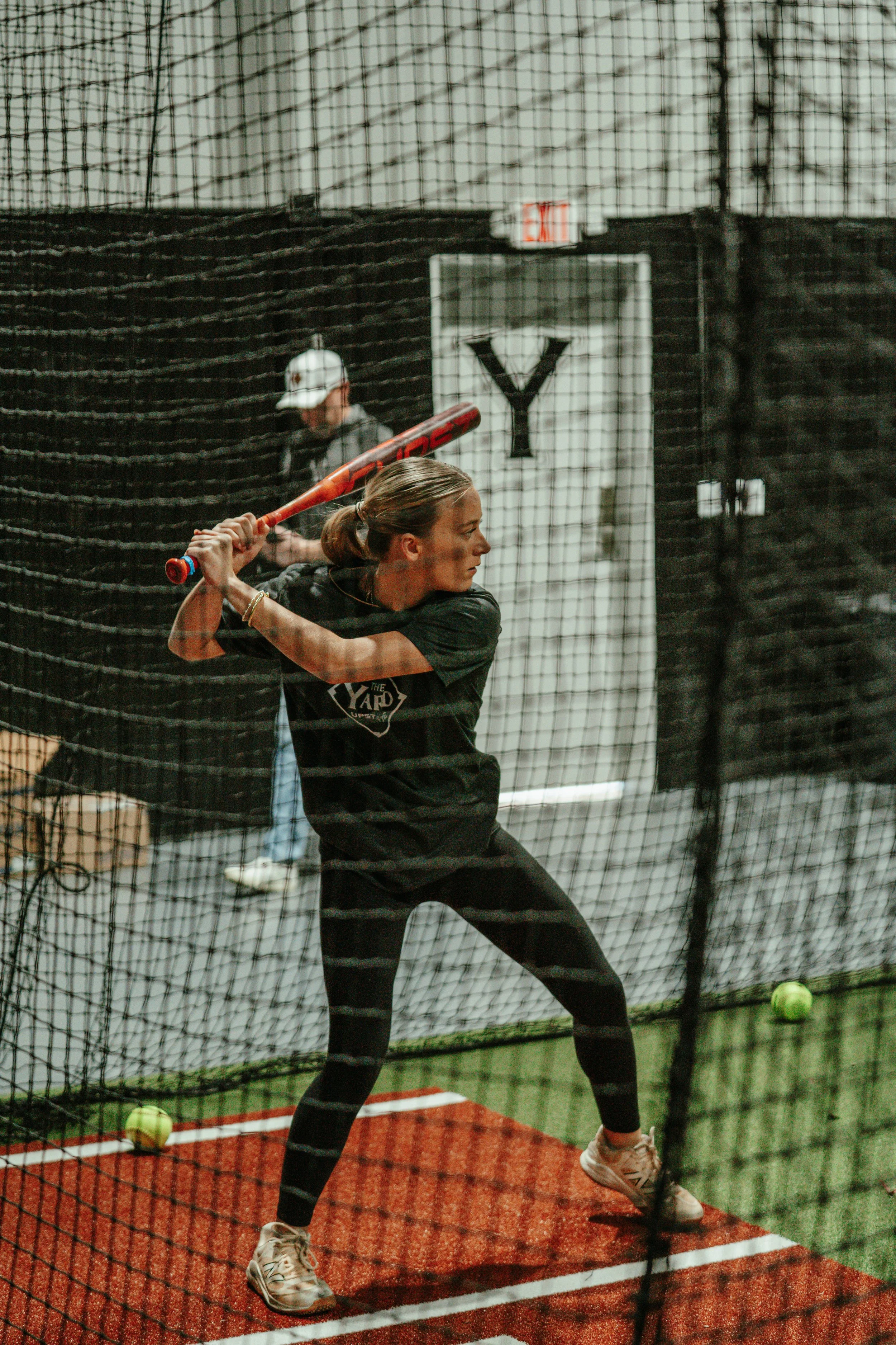 A girl practicing sofball in an indoor batting cage called The Yard Upstate, holding a bat and preparing to swing.