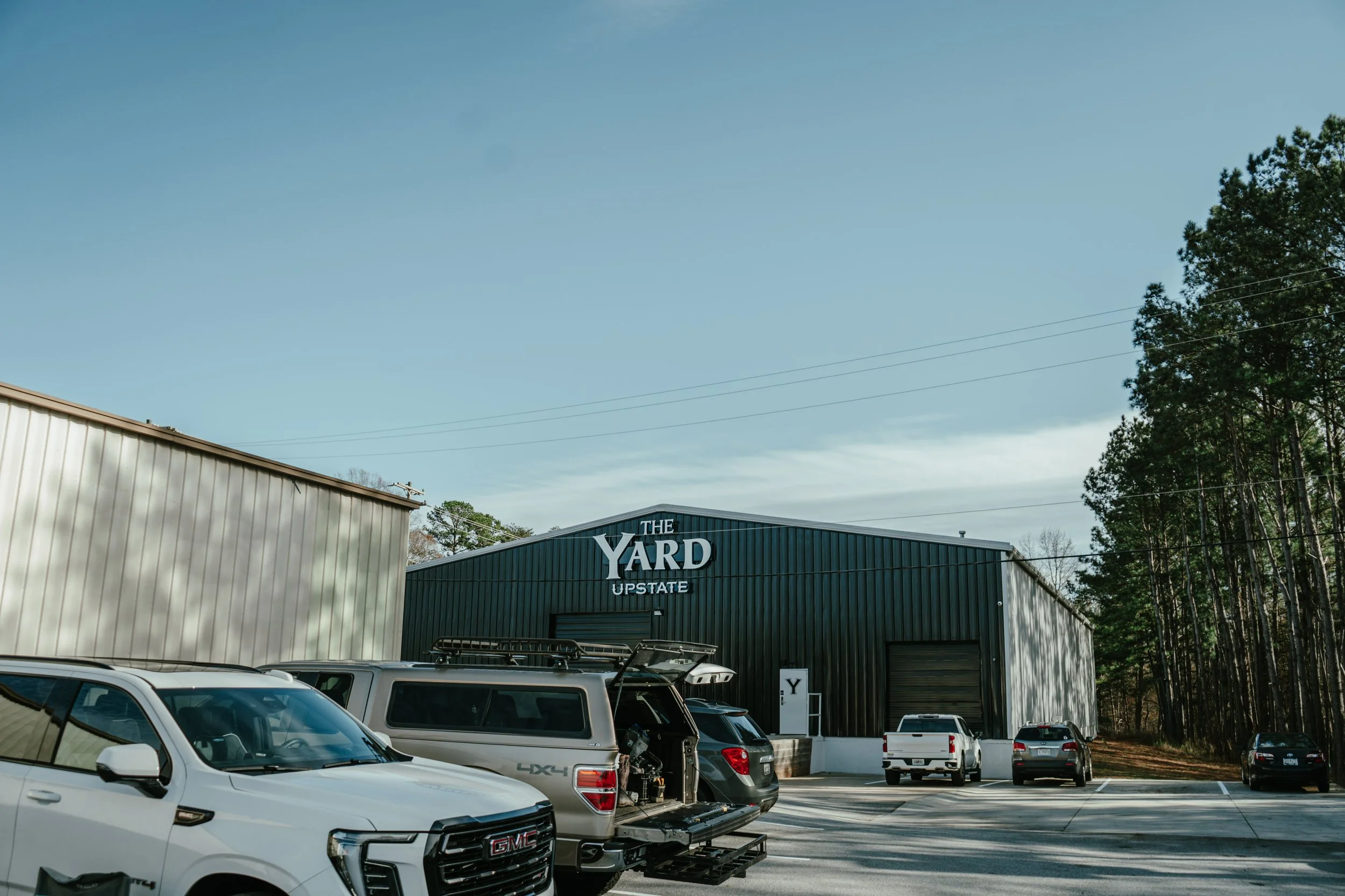 Exterior view of a large black industrial building with a sign that reads 'The Yard Upstate'. Several parked cars are visible in the parking lot, with trees lining the background on the right side.