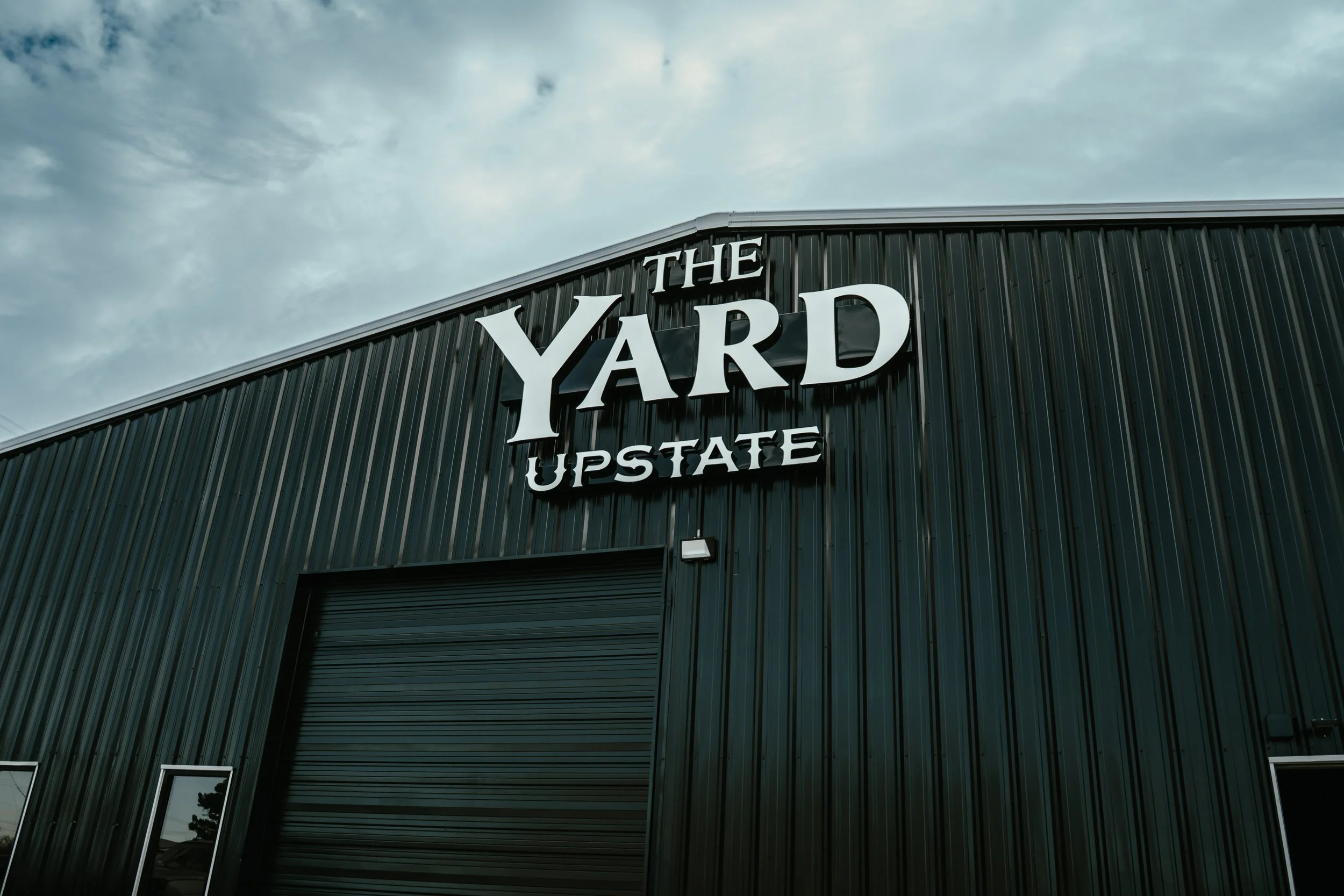 A large dark green metal building with a sign that reads 'The Yard Upsate' on the front, under a cloudy sky.