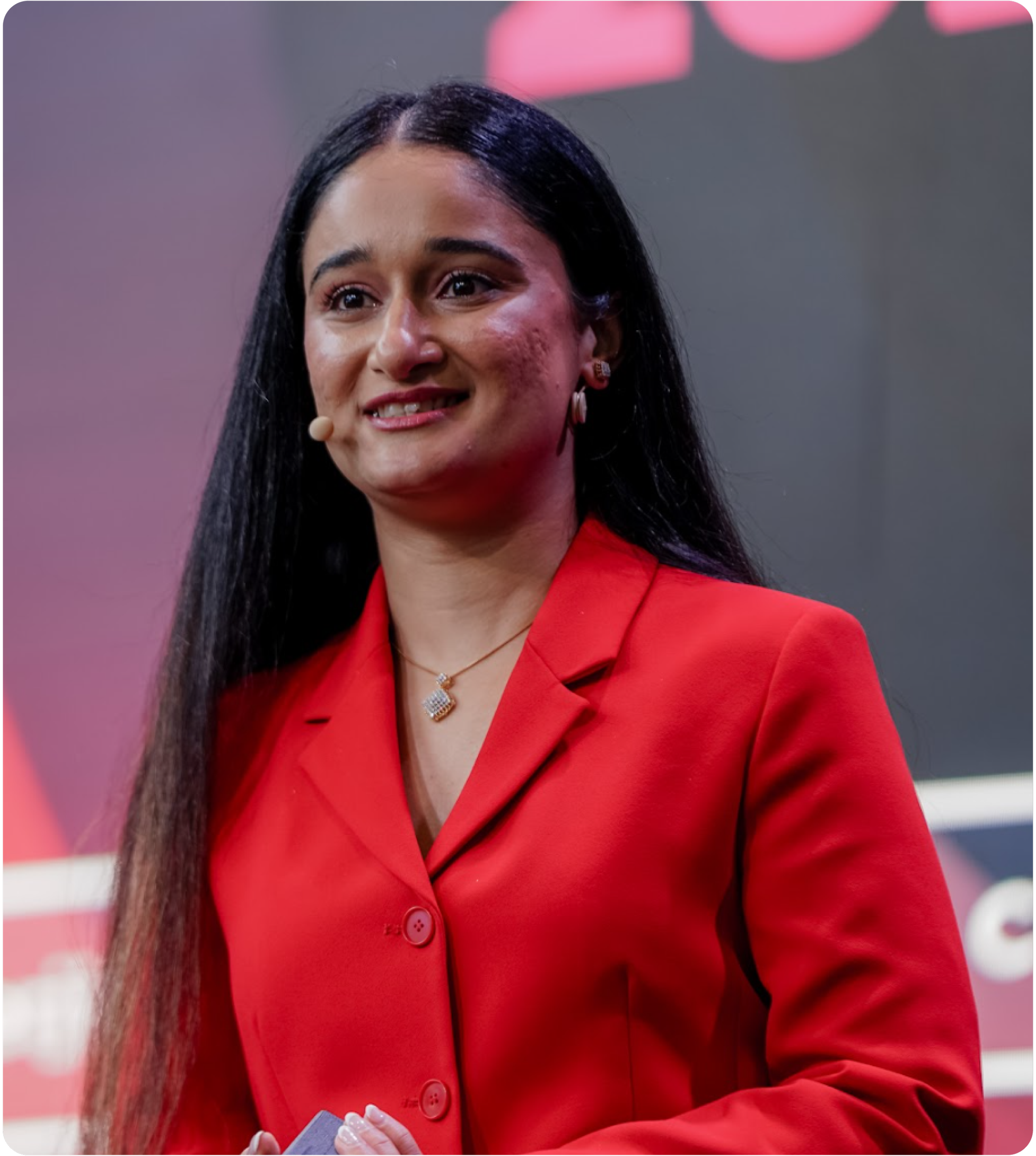 Jeet Bhatti with long black hair, wearing a red blazer and jewelry, speaking on stage.