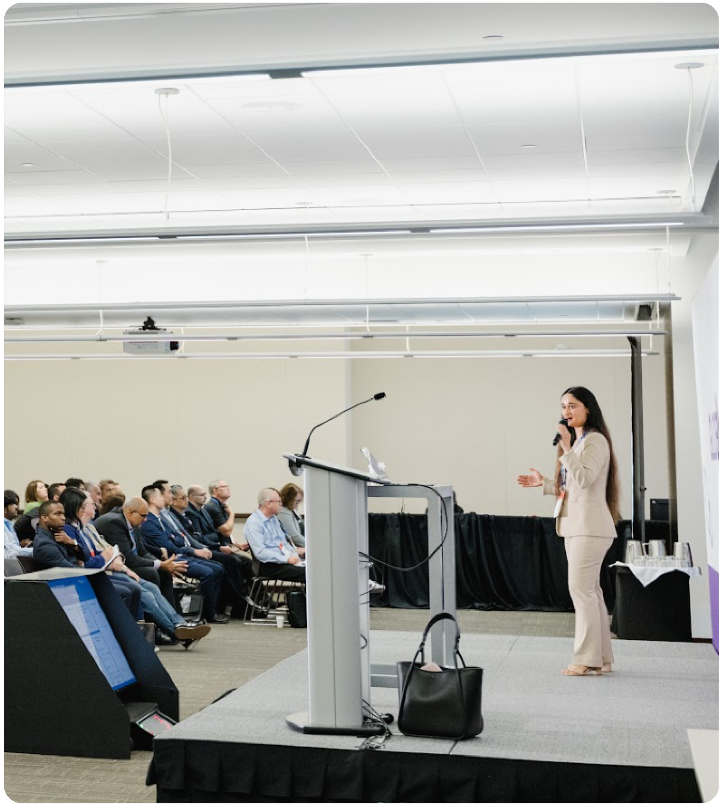 A woman in a beige suit giving a presentation to an audience at a conference. She is standing next to a podium with a microphone and a laptop, holding a microphone in her right hand, explaining with her left hand. The audience, seated in rows, is watching her attentively in a large, well-lit room with a high ceiling.