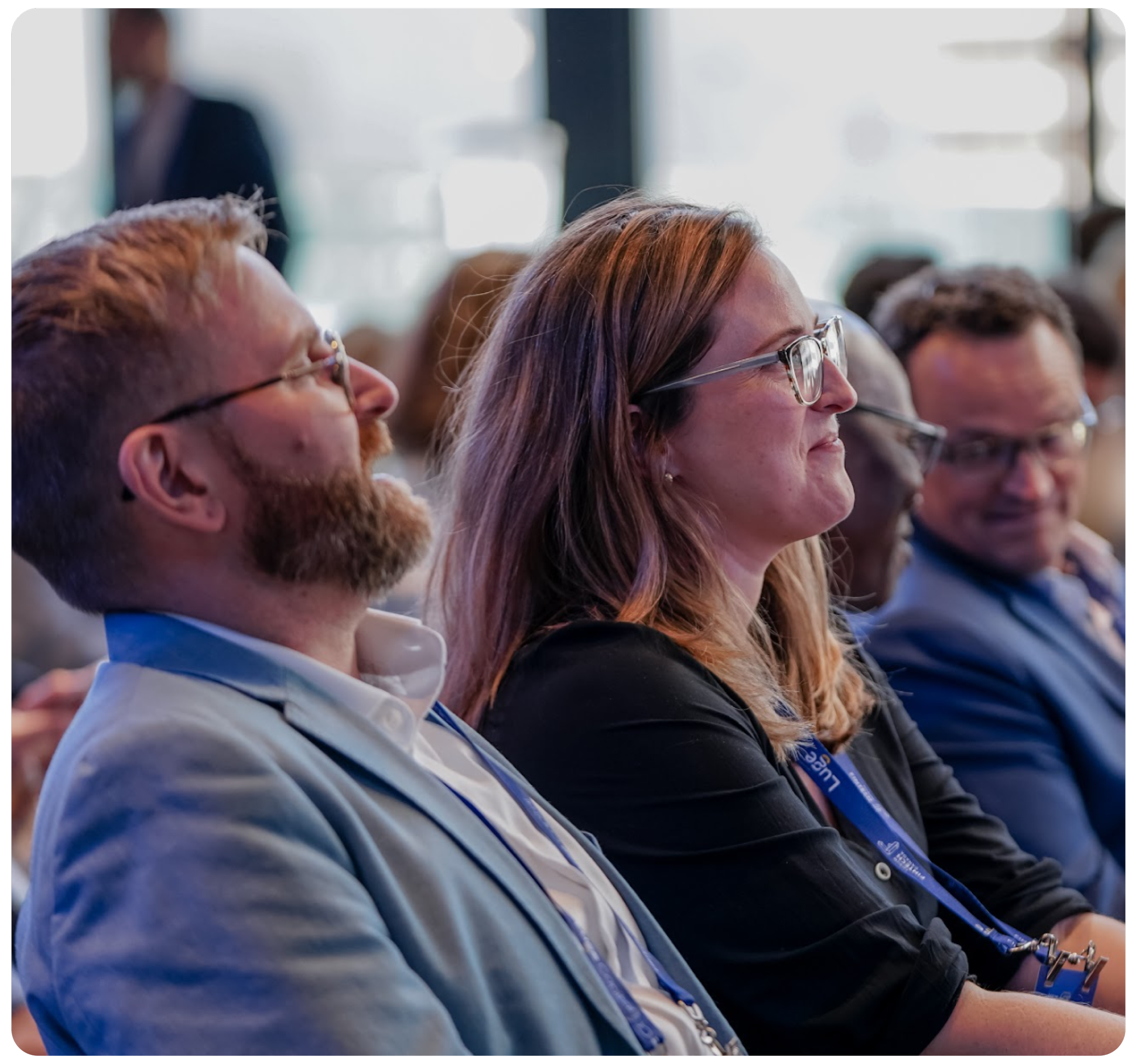 People attending a conference, seated and listening attentively, wearing conference badges, with some smiling.