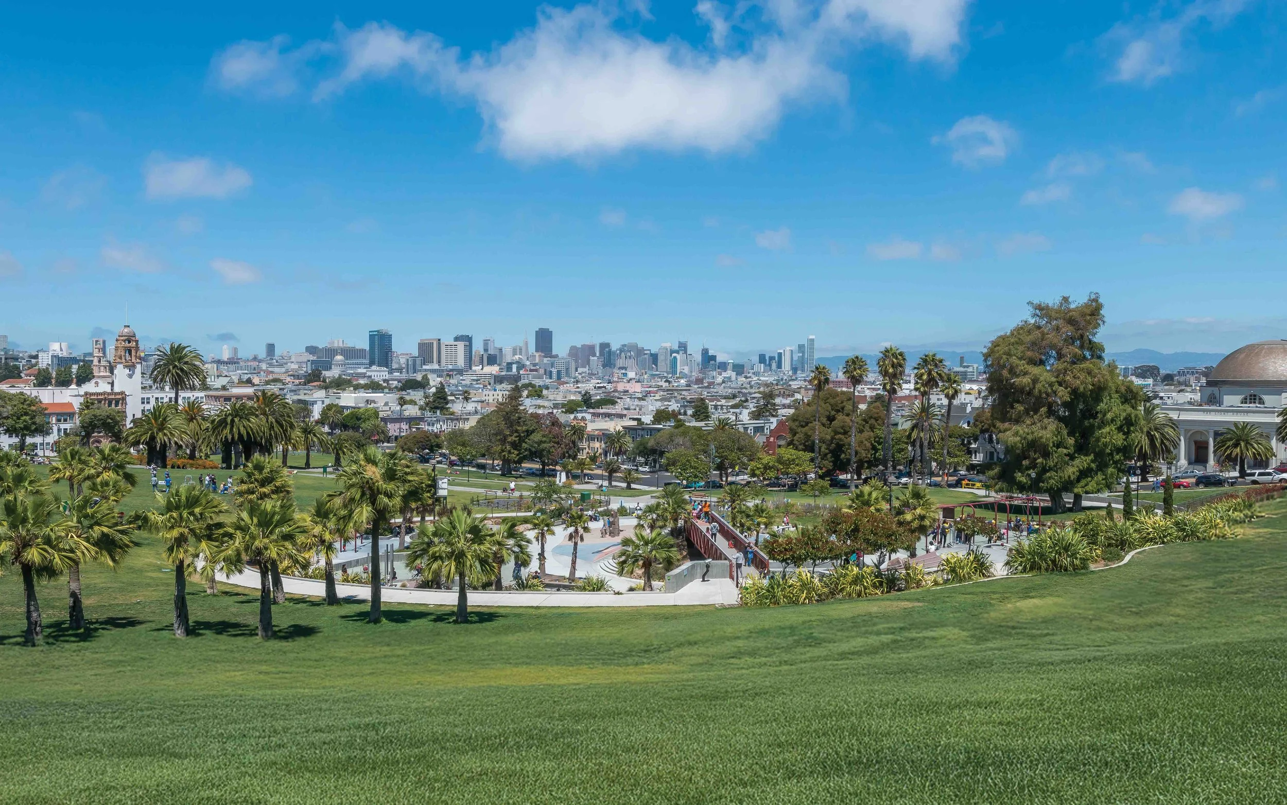 Views of a city skyline with tall buildings in the distance, lush green park in the foreground with palm trees, walkways, and a few people enjoying the area on a sunny day.