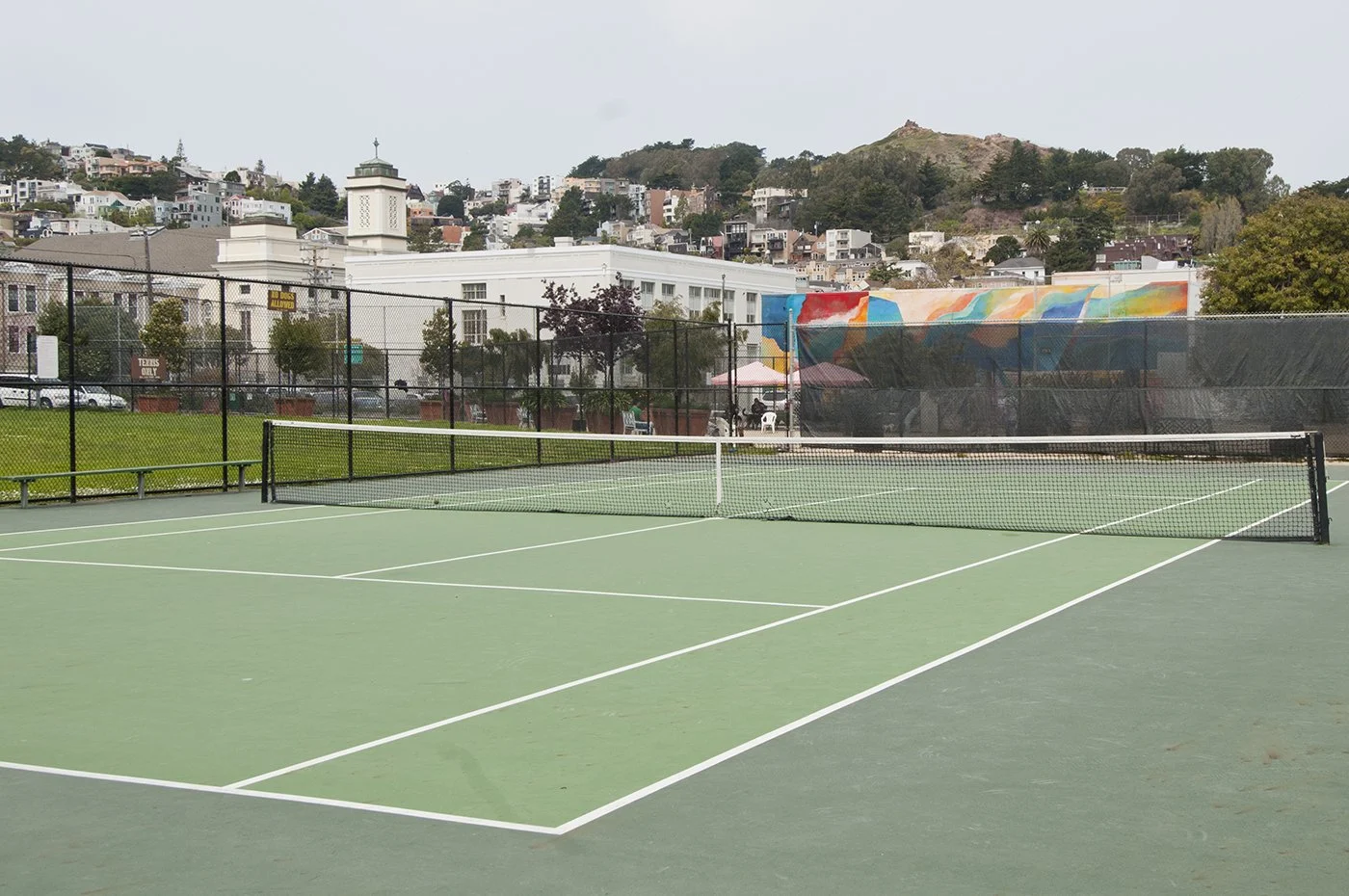 Empty tennis court with a net, surrounded by a fence, with a cityscape and hillside in the background.