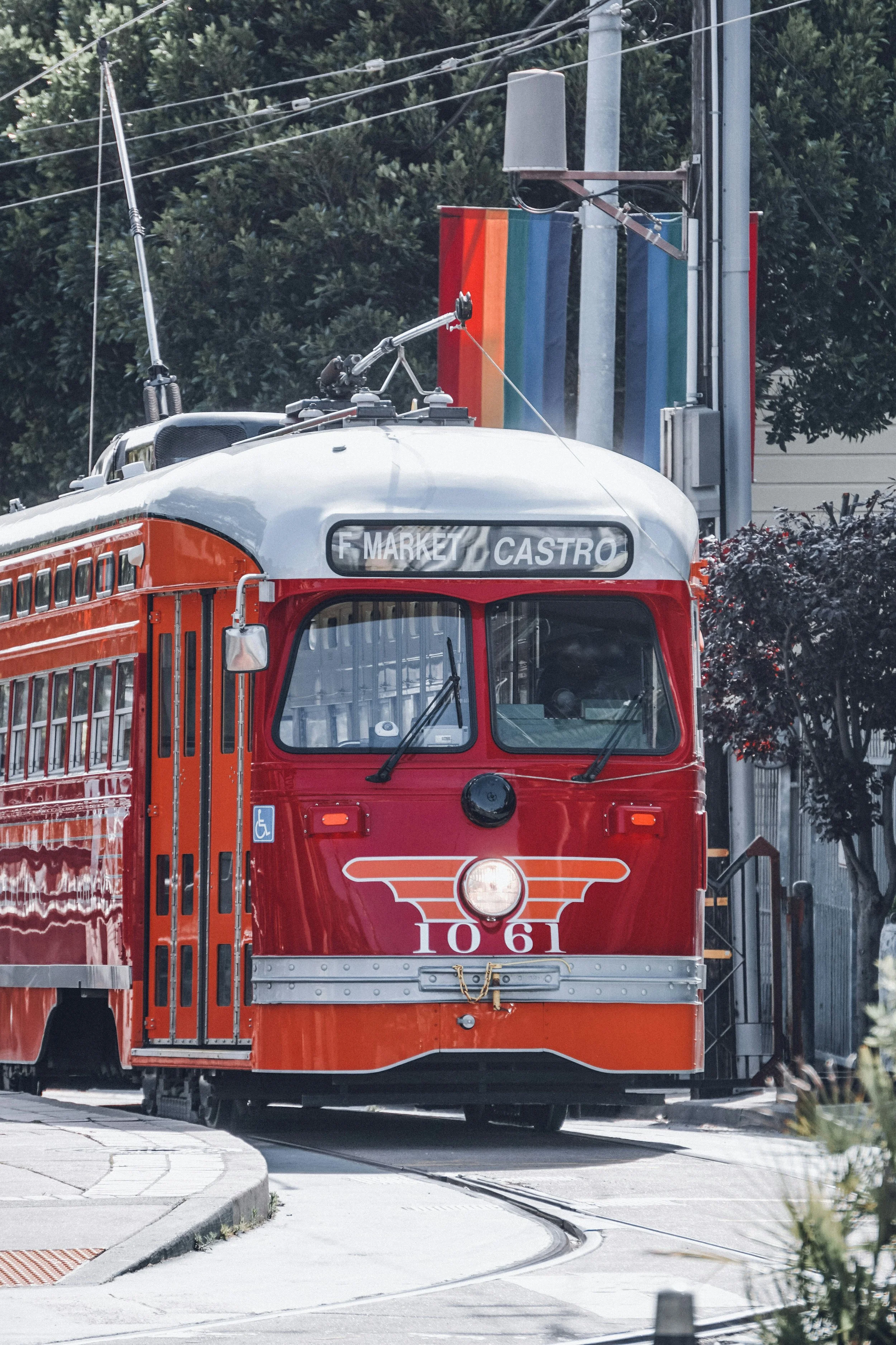 A vintage red streetcar on tracks, with a destination sign indicating 'F. Market' and 'Castro', moving through an urban area with trees and power lines.