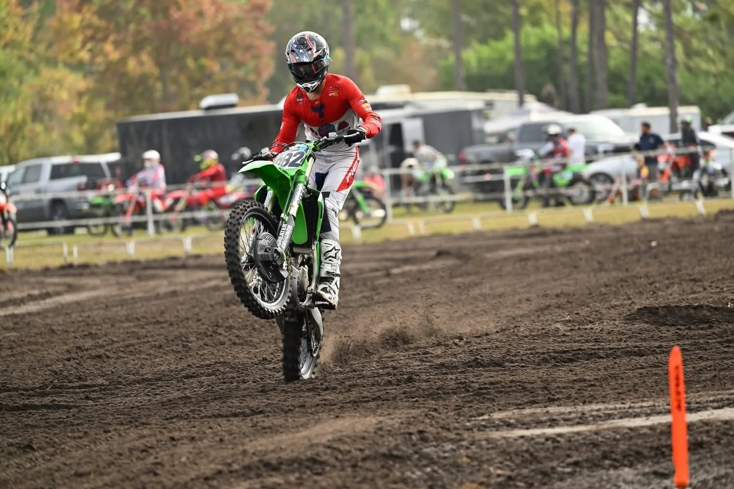A motocross rider in a red and white outfit performing a wheelie on a green dirt bike on a dirt track, with cars and spectators in the background.