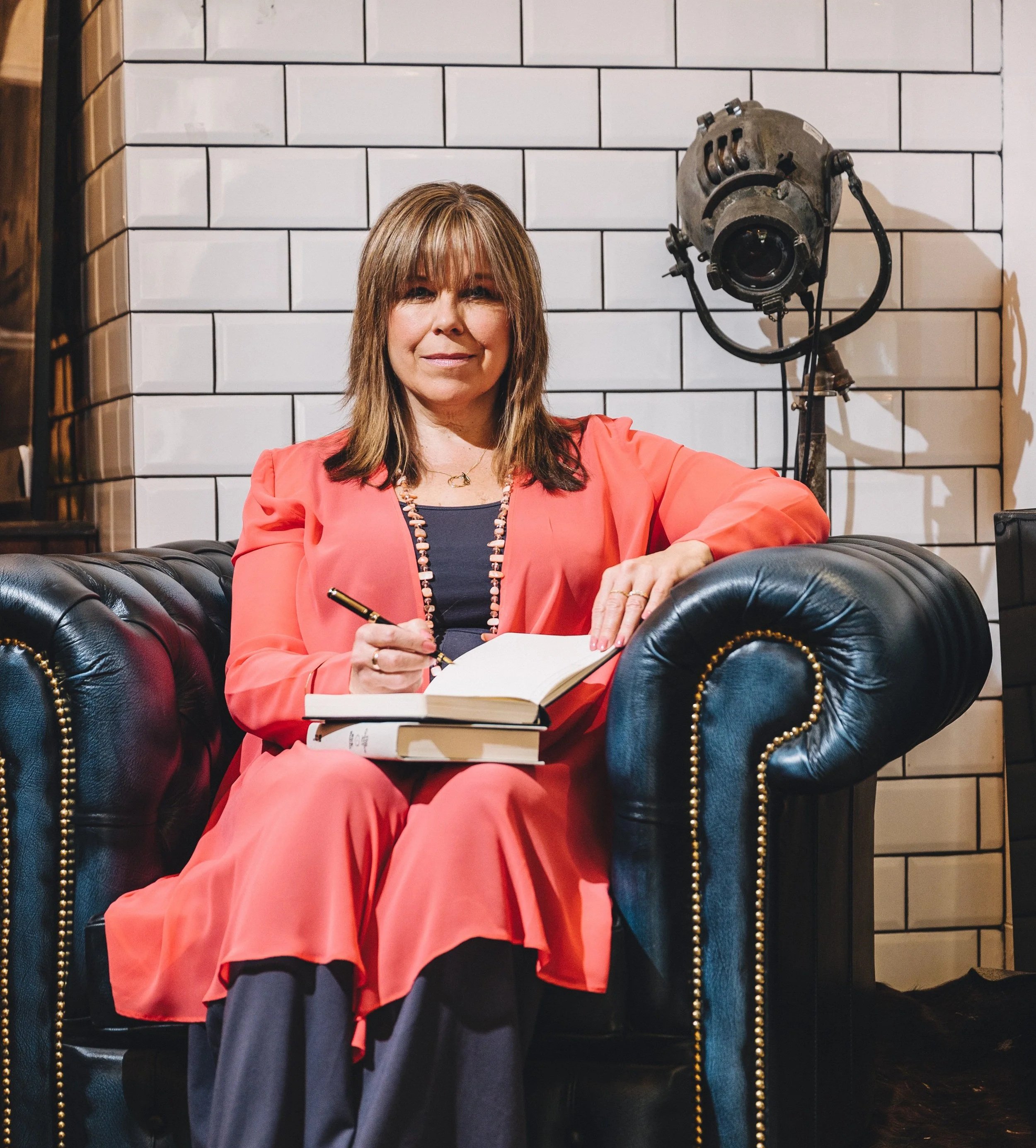 Fiona sitting in a navy leather armchair with a note book looking into the camera