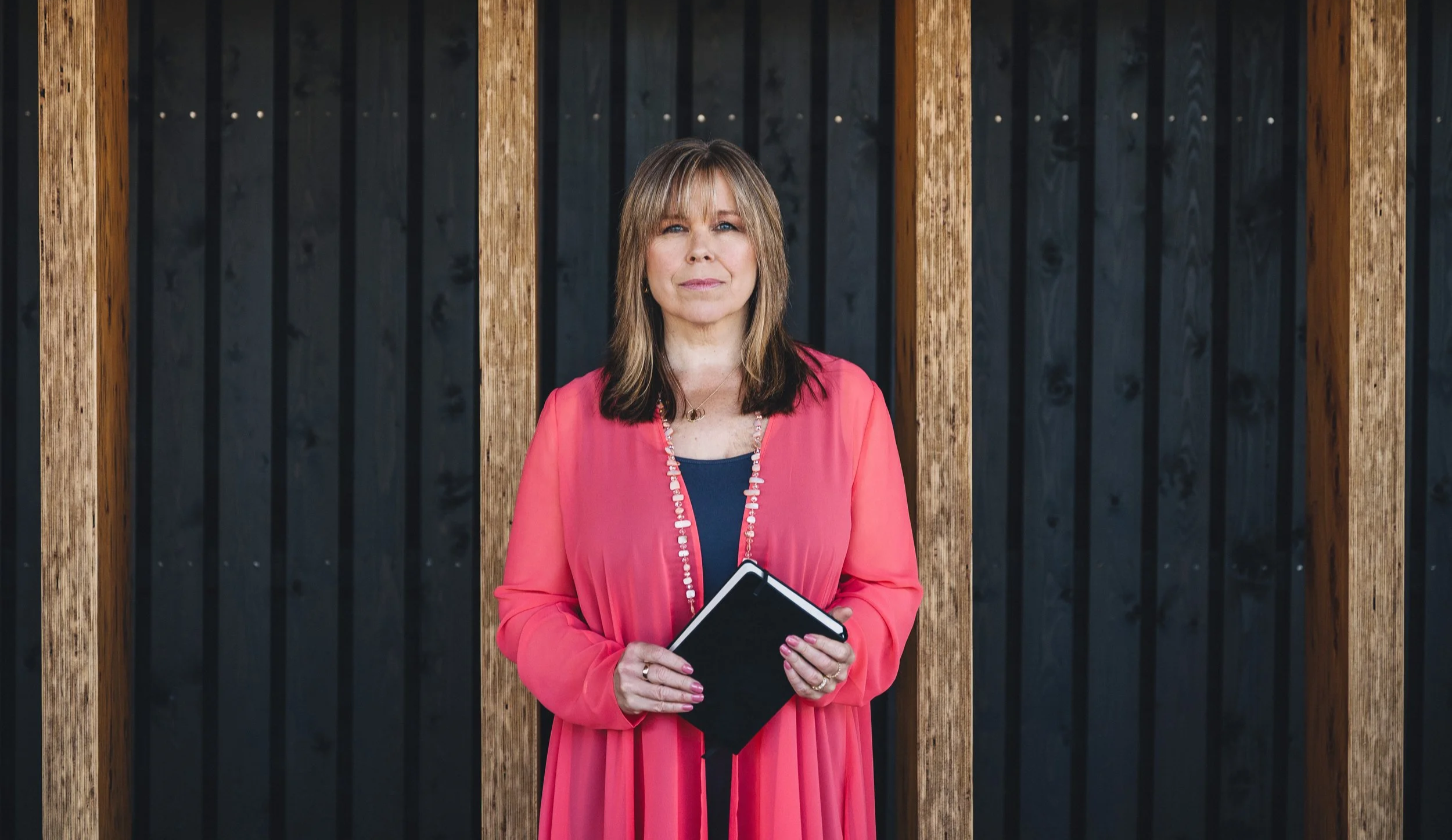 A woman standing in front of a background of wooden beams and dark vertical panels, holding a black folder, wearing a coral pink dress with a beaded necklace.