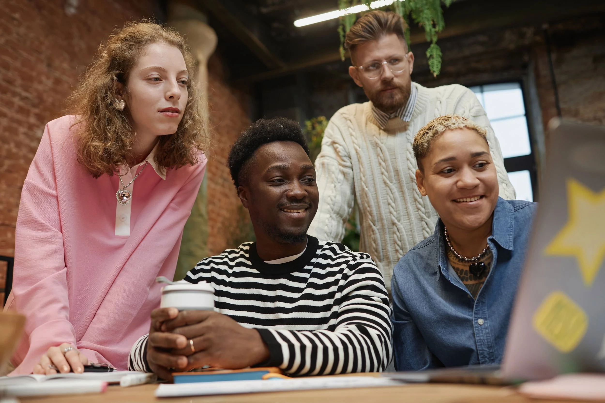 Five diverse people gathered around a laptop, looking at the screen with smiles, in a cozy brick-walled room with plants and natural light.