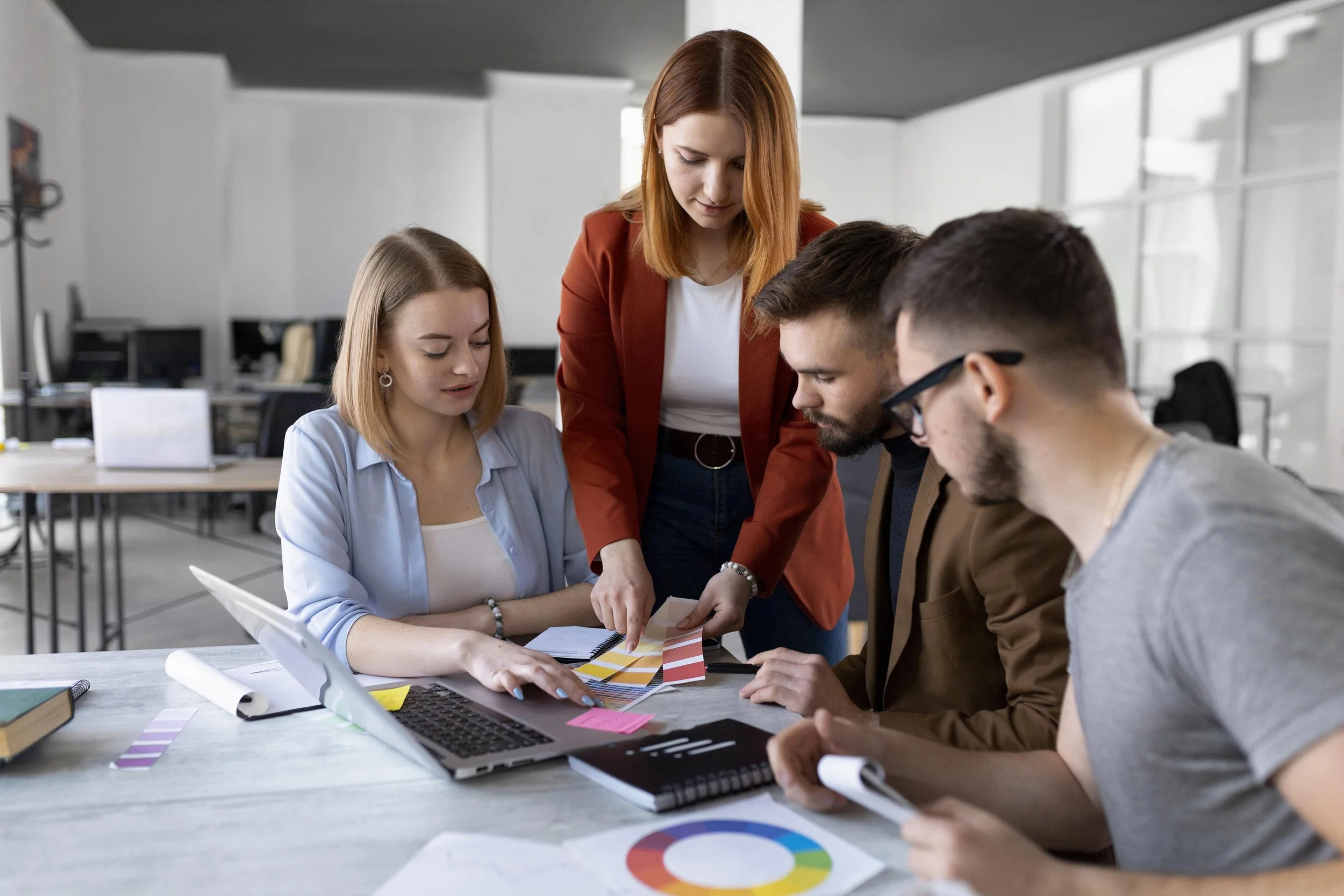 Group of four young adults collaborating around a table in a modern office, reviewing materials and a laptop.