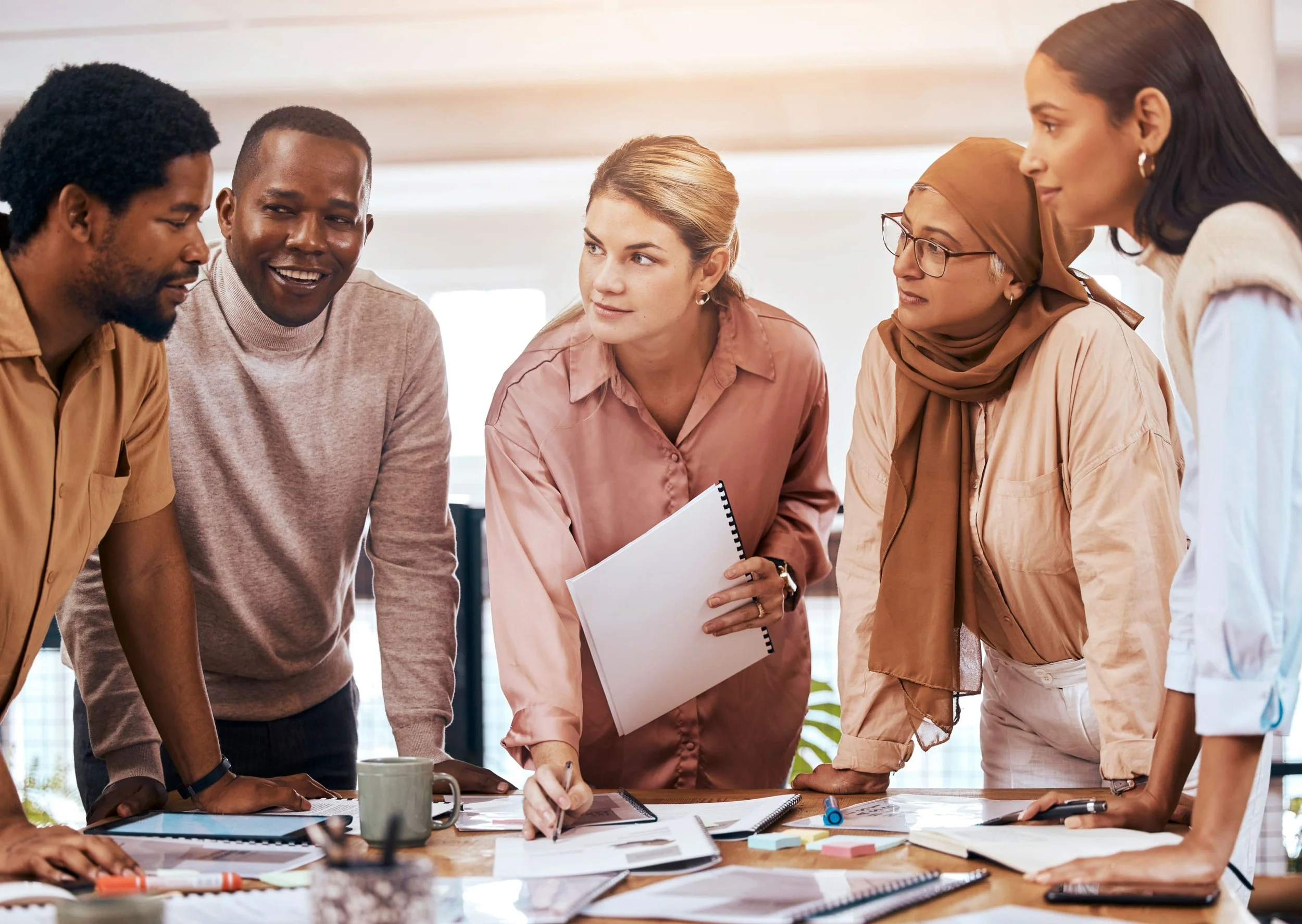 A diverse group of five people gathered around a table, discussing documents and working together in an office setting.