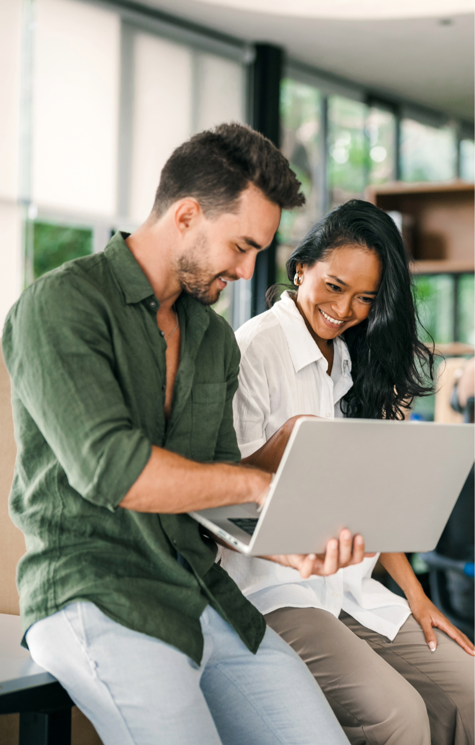 A man and woman sitting together, looking at a laptop and smiling, in a bright, modern indoor space.