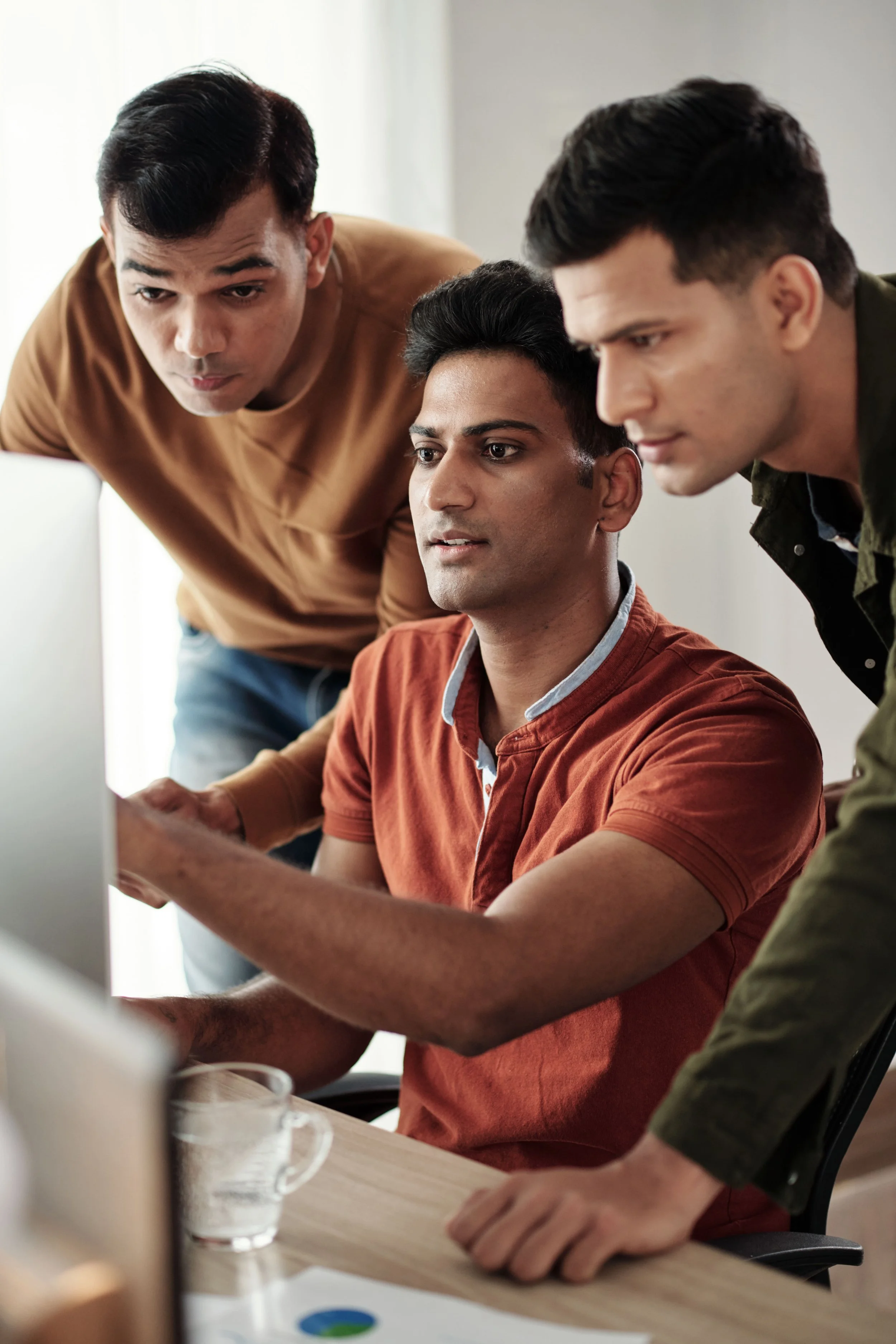 Three men gathered around a computer, focused and collaborating on a project in an office setting.