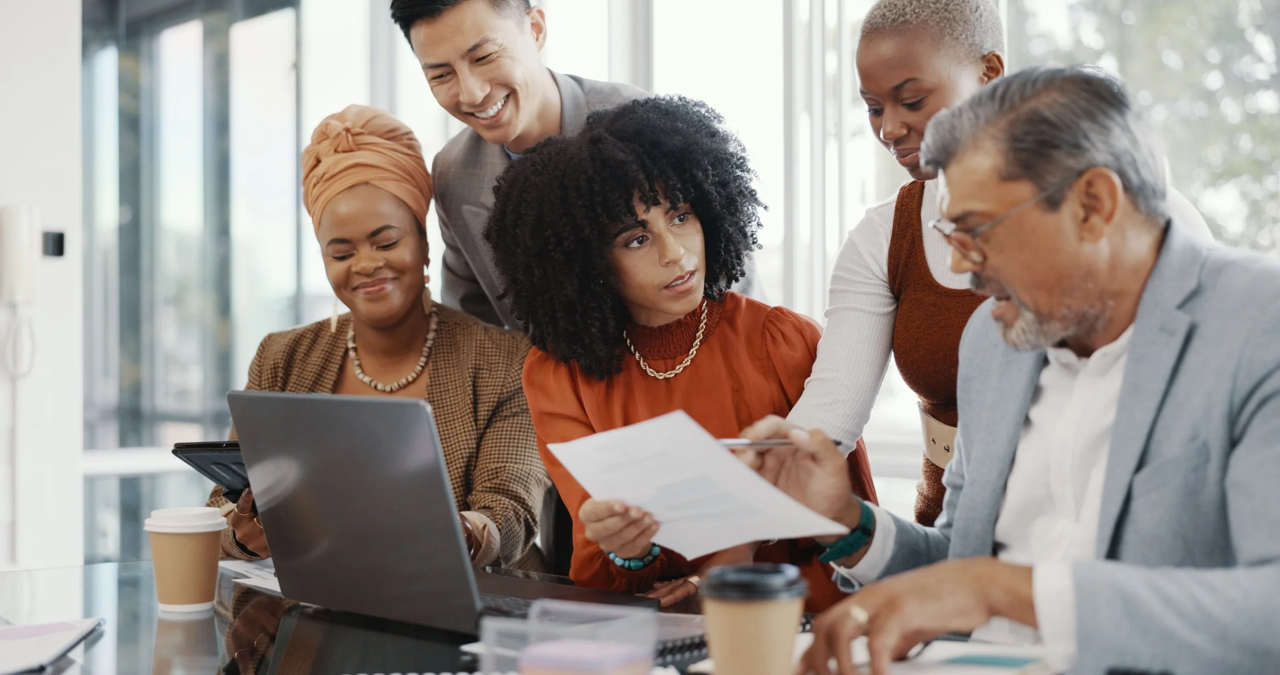 Group of five diverse professionals working together in a modern office, looking at documents and a laptop, with coffee cups on the table.