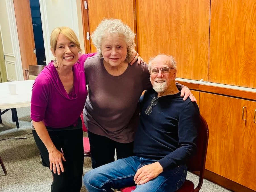Three people smiling and posing together in a room with wooden cabinets in the background. One woman is wearing a magenta top, another woman is wearing a dark shirt, and a man is seated wearing glasses and a black shirt.