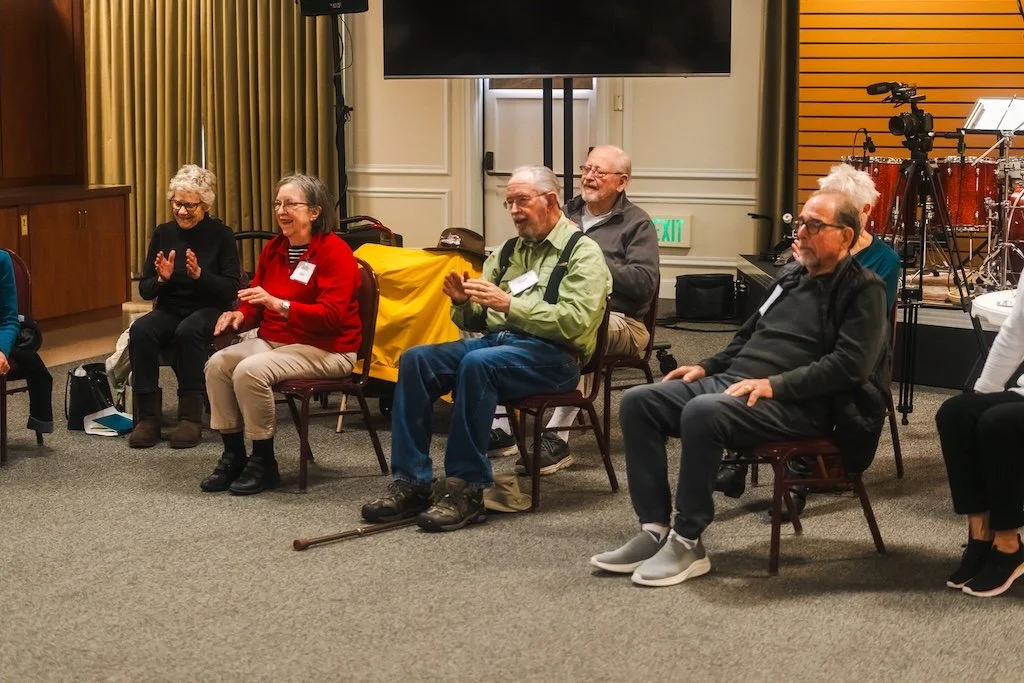 Group of elderly people sitting on chairs in a room, some smiling and clapping, with musical instruments and a camera on a tripod in the background.