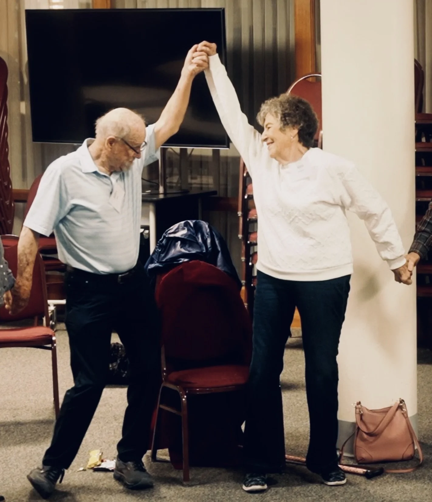 An elderly man and woman smiling as they dance together, holding hands in a circle, in a room with chairs and a large television in the background.