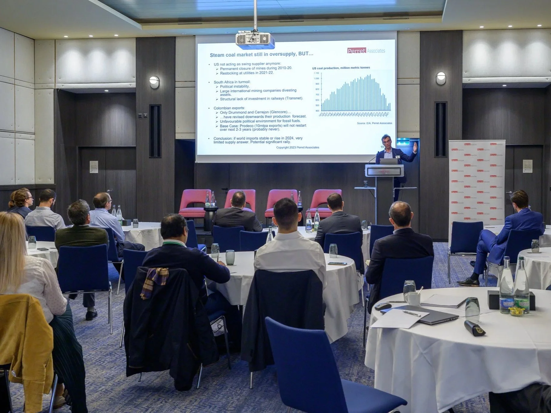 A conference room with attendees listening to a presenter at the front. The presenter is standing behind a podium, pointing to a large screen displaying a presentation slide. The audience is seated at round tables with notebooks and bottles of water.