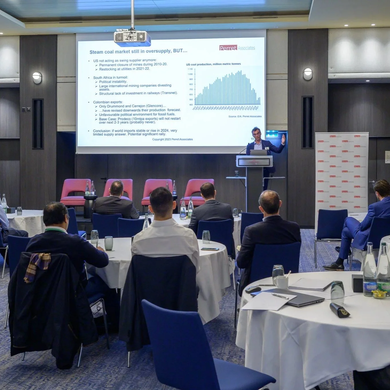 A man giving a presentation at a conference room with a large screen displaying a PowerPoint slide titled 'Steam coal market still in oversupply, BUT...'. The slide includes bullet points, a graph of US coal production in million metric tonnes, and the Perret Associates logo. The audience, dressed in formal attire, is seated around round tables with notebooks, water bottles, and glasses.
