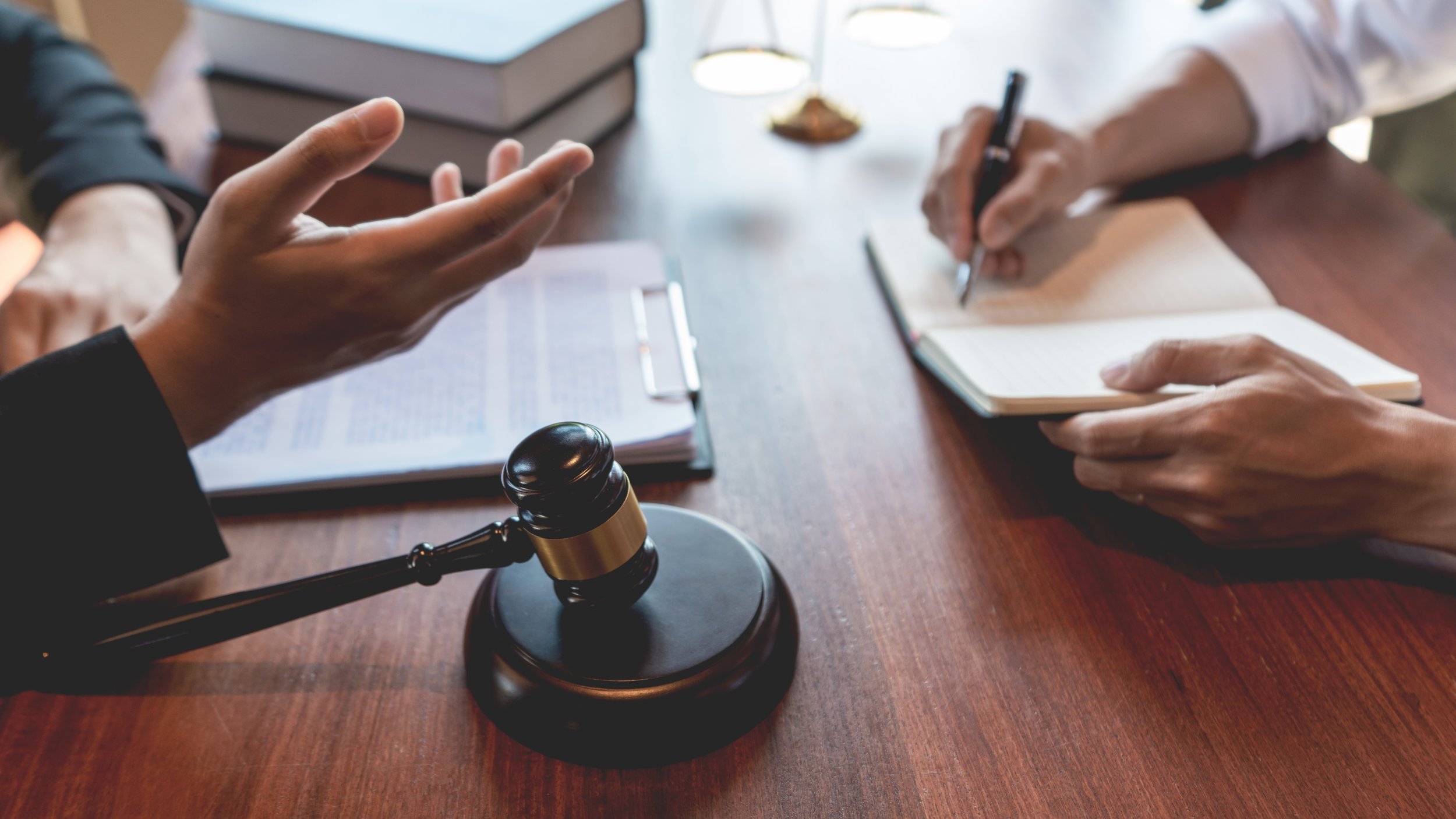 A judge's gavel on a wooden table, with two people discussing a legal matter; one person is gesturing with their hand, and the other is writing in a notebook.