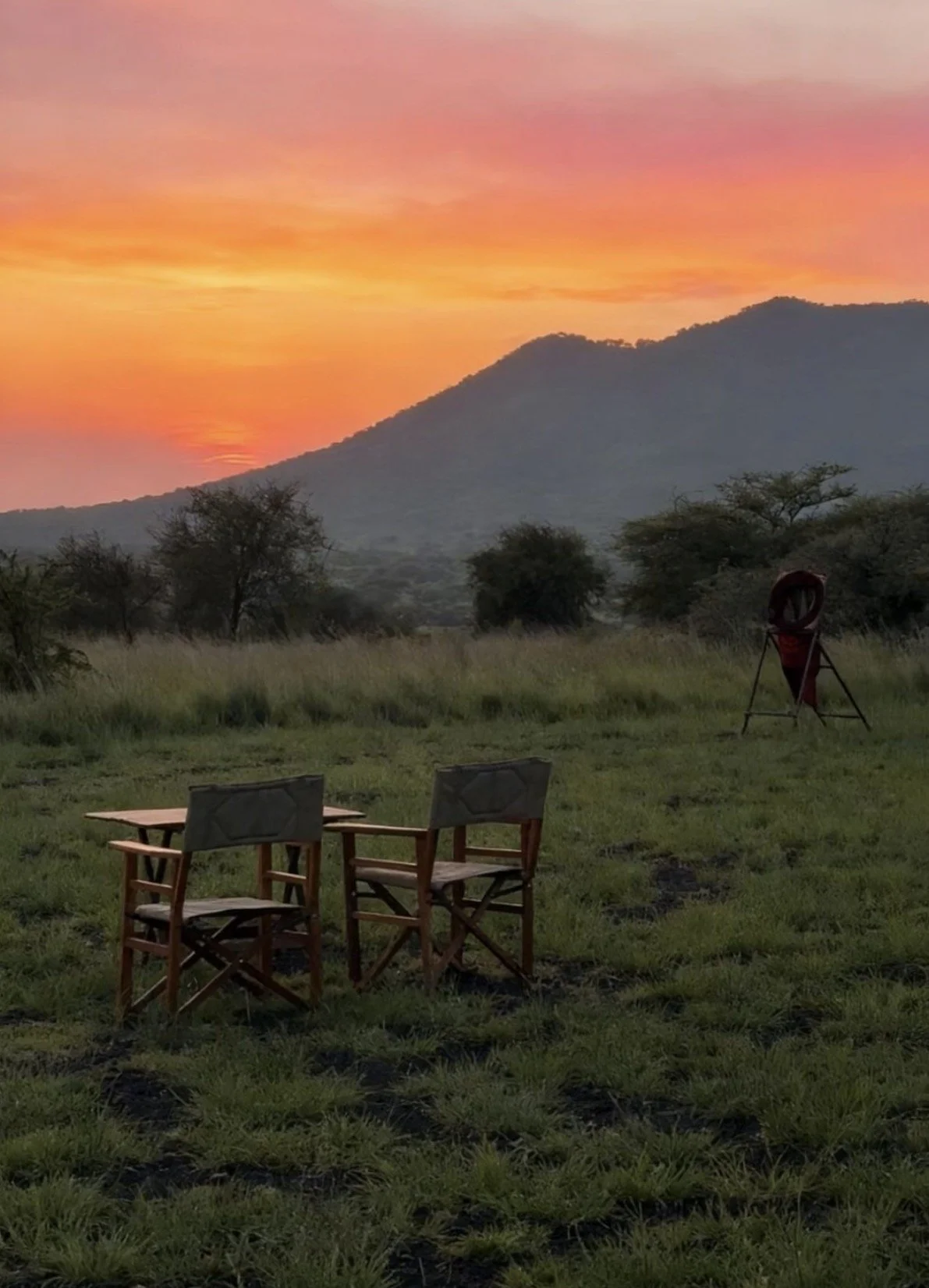 A scenic view of a sunset over mountains with a grassy field in the foreground in Tsavo West national park, East AFrica There are two wooden chairs and a table, along with a piece of camping or outdoor equipment.