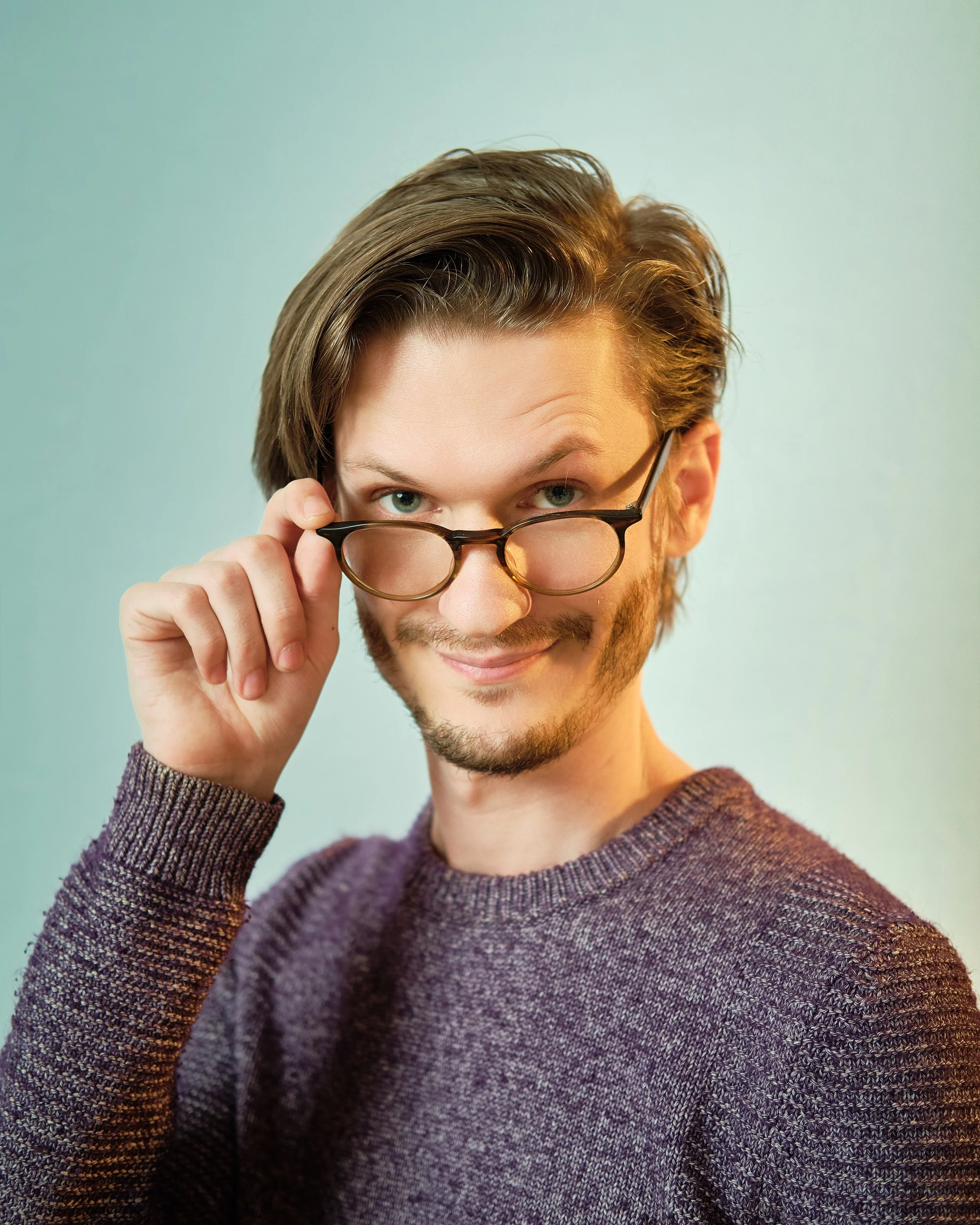 A man with light skin, brown hair, and a beard wearing a purple sweater, holding glasses and looking directly at the camera with a confident expression.