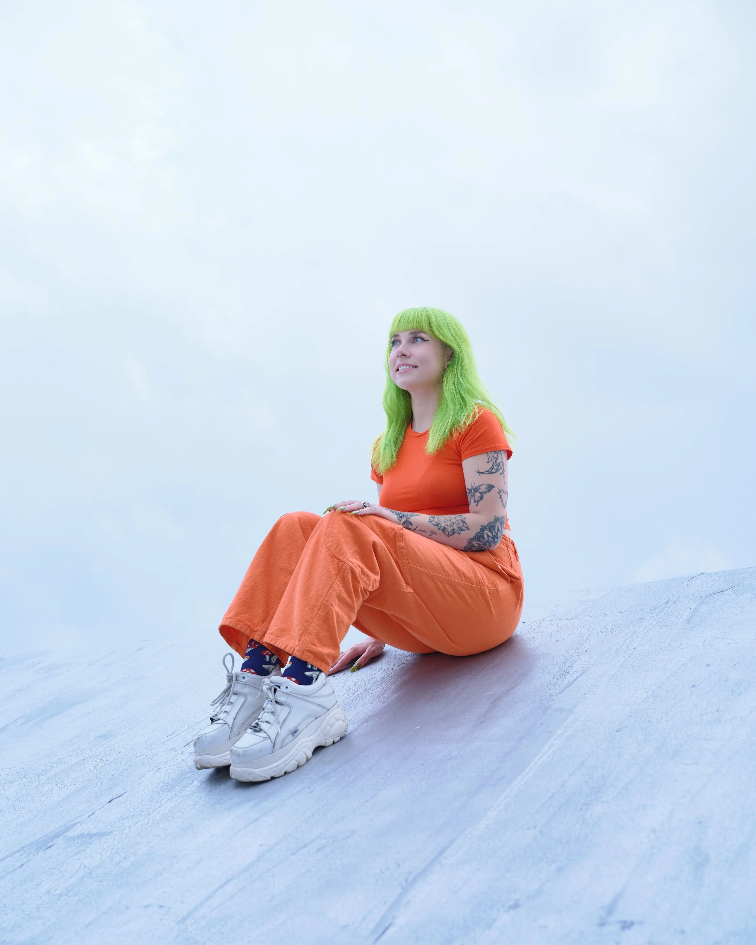 A woman with bright green hair sitting on a white surface, wearing an orange outfit and white sneakers, with a cloudy sky in the background.