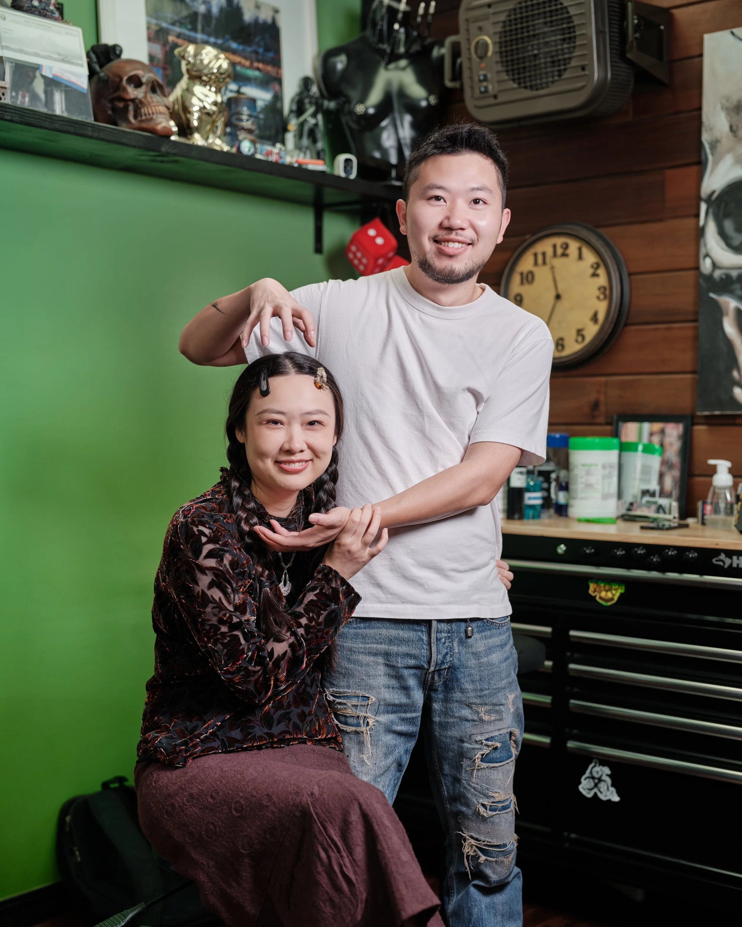A man standing behind a seated woman, styling her hair with clips in a room with green and wooden walls, decorated with various objects, including a clock and skulls.