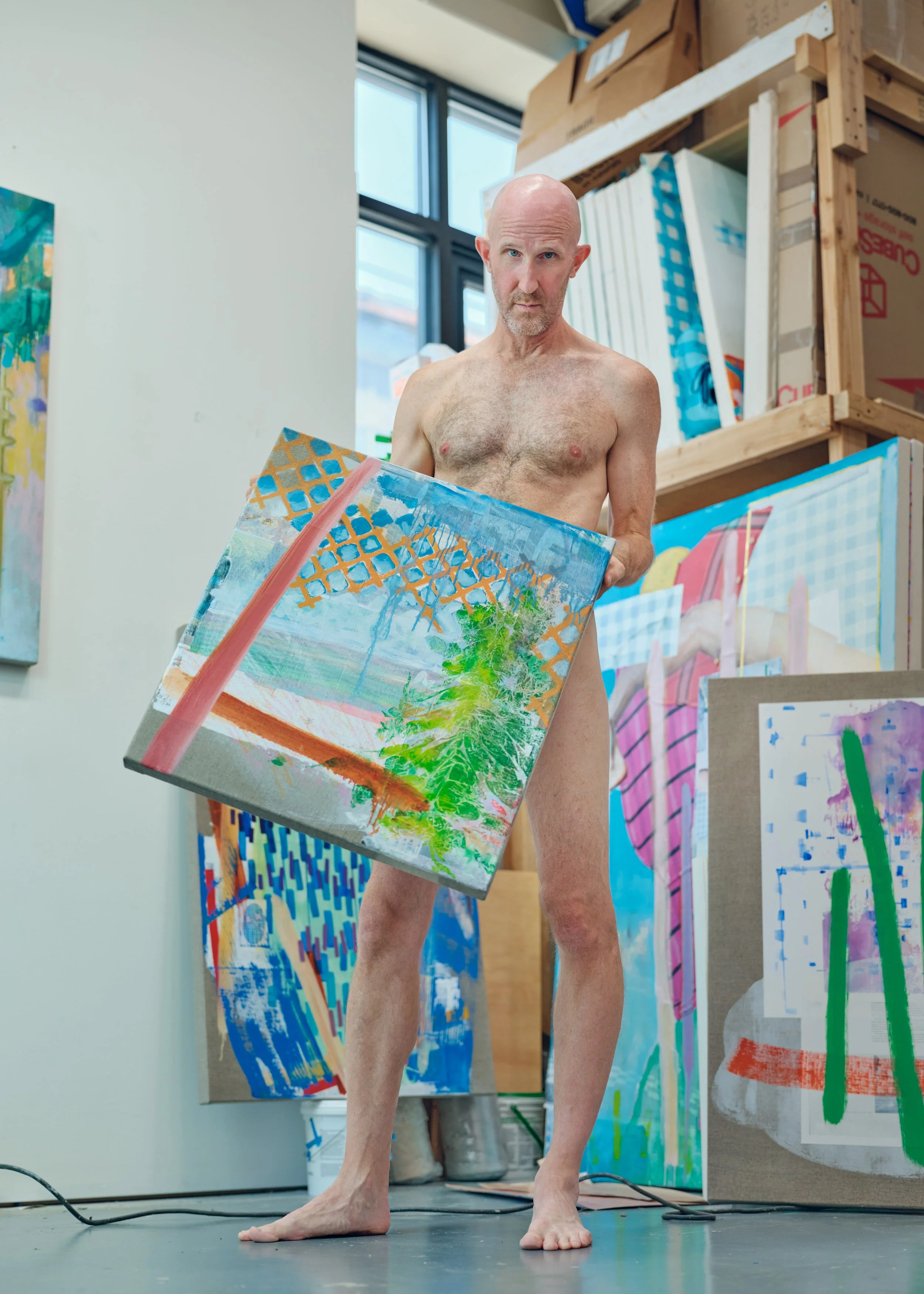 A shirtless man with a bald head and a beard standing in an art studio, holding a colorful abstract painting. The studio has windows, shelves with art supplies, and several other colorful paintings in the background.
