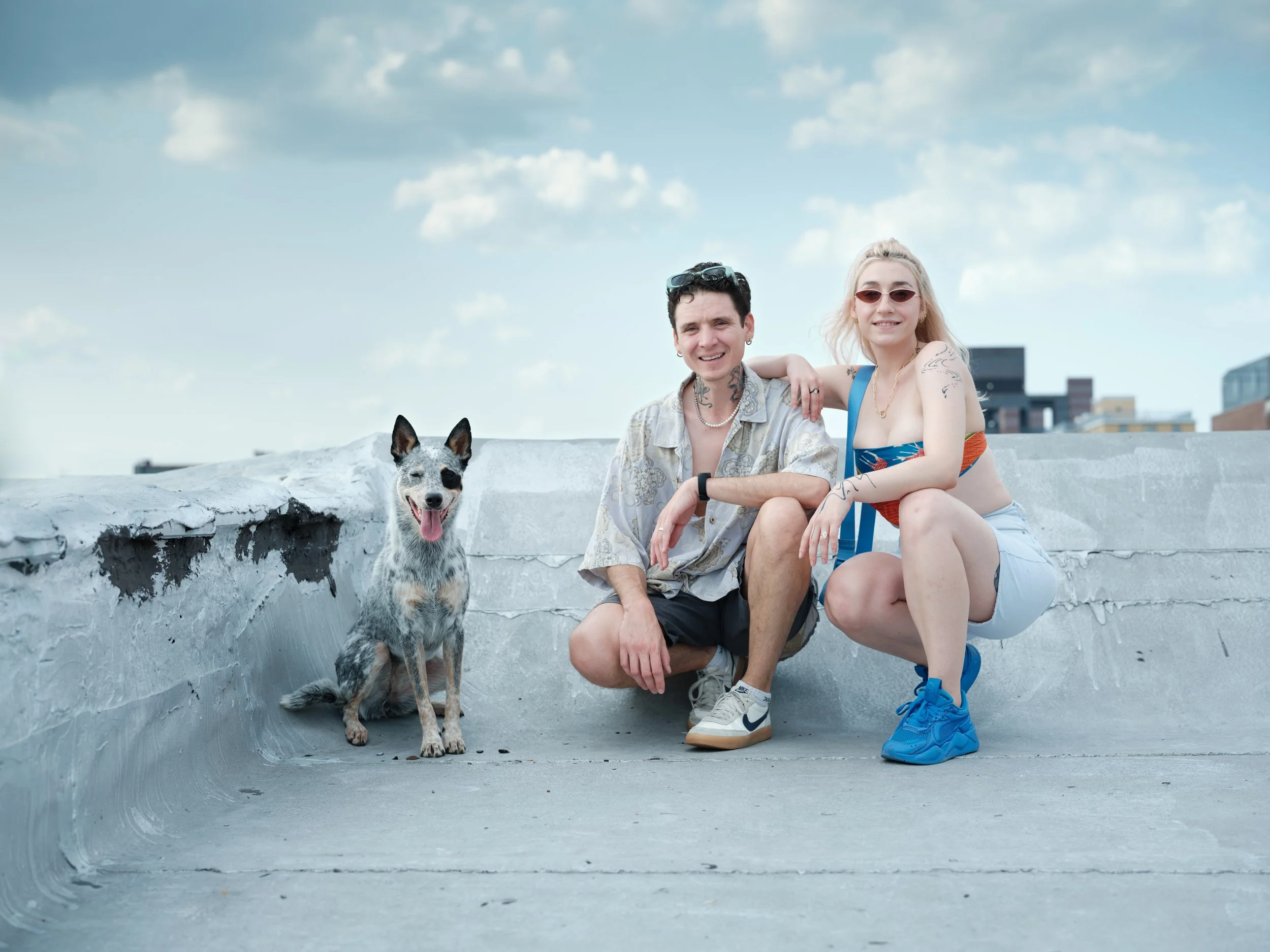 A young man, a young woman, and a dog sitting on the edge of a concrete surface outdoors on a sunny day.