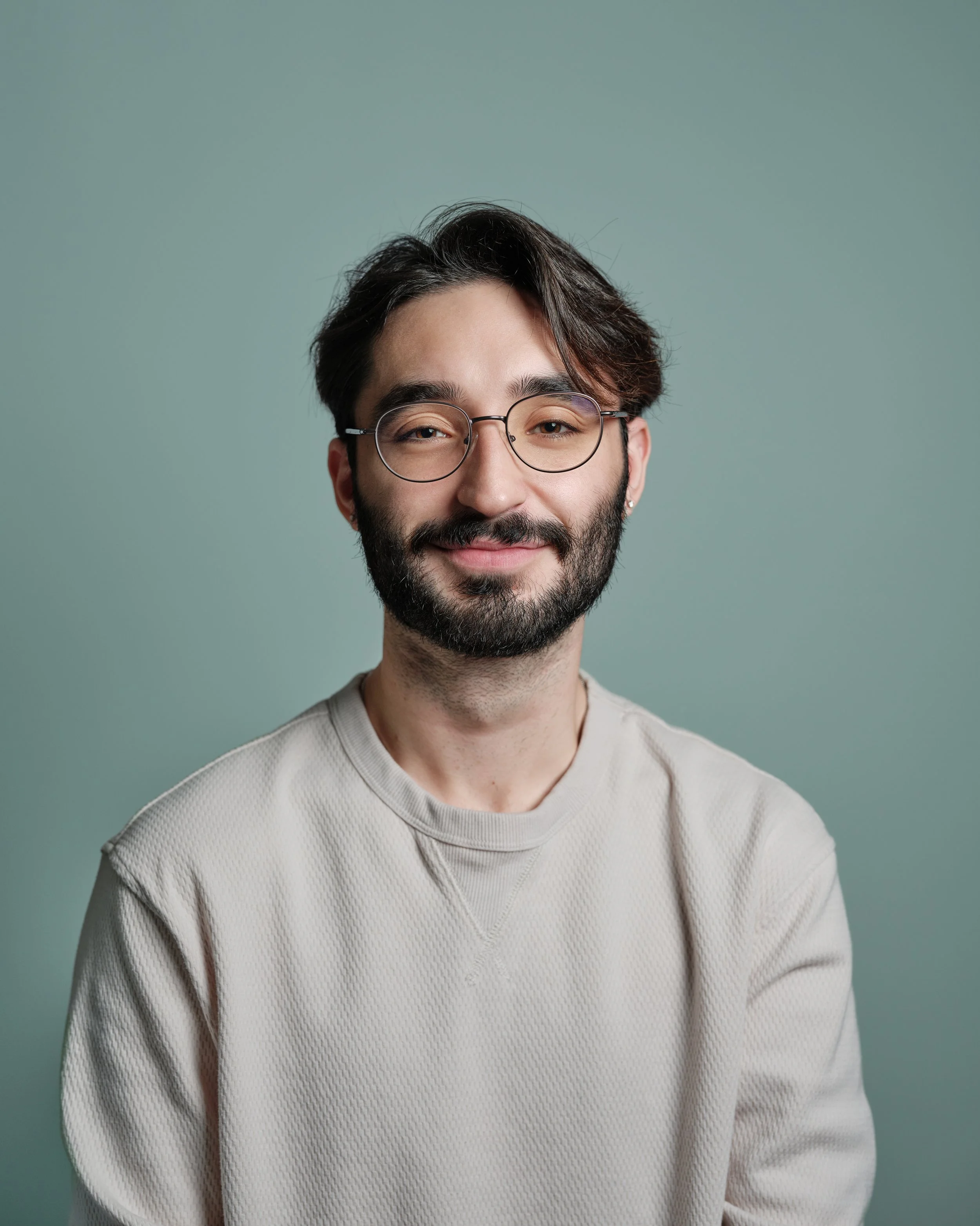 Headshot of a smiling man with dark hair, glasses, and a beard, wearing a light-colored sweatshirt, against a plain green background.