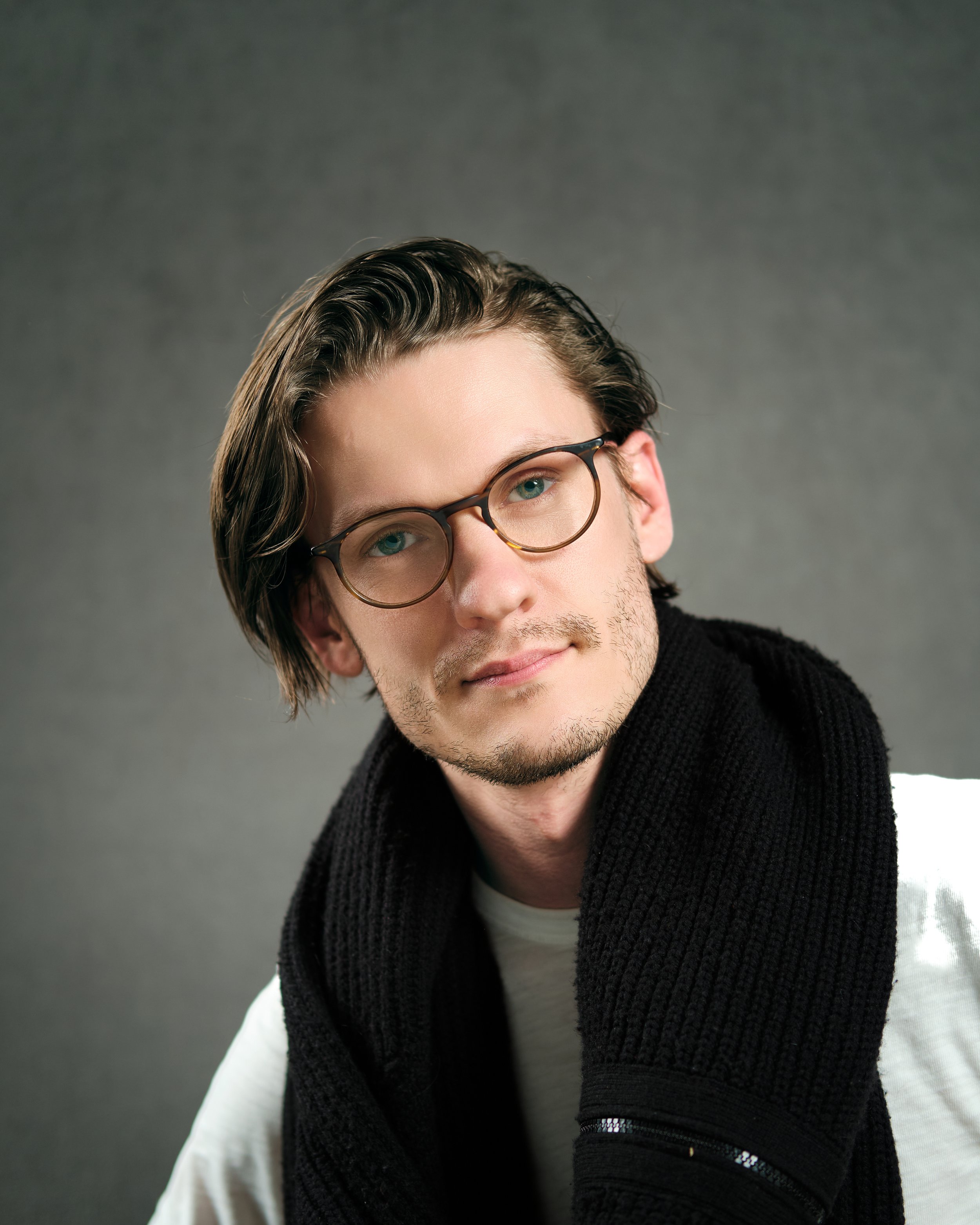 Portrait of a young man with medium-length brown hair, wearing glasses, a white shirt, and a black knitted scarf, against a gray background.