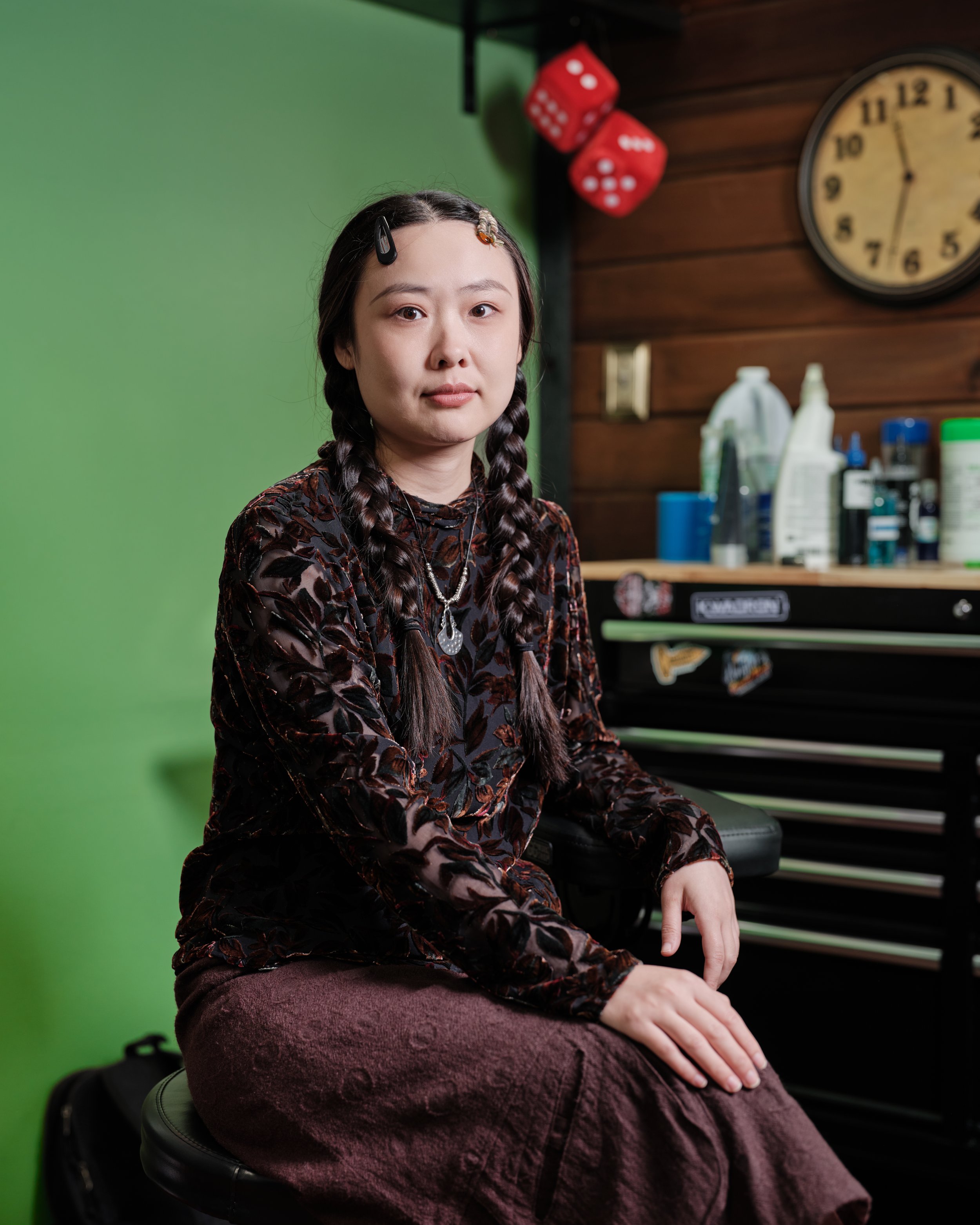 A young woman with braided hair, wearing a dark patterned top, sits on a black stool in a tattoo studio, with tattoo inks and supplies behind her, a green screen wall to her left, and tattoos visible on her neck.