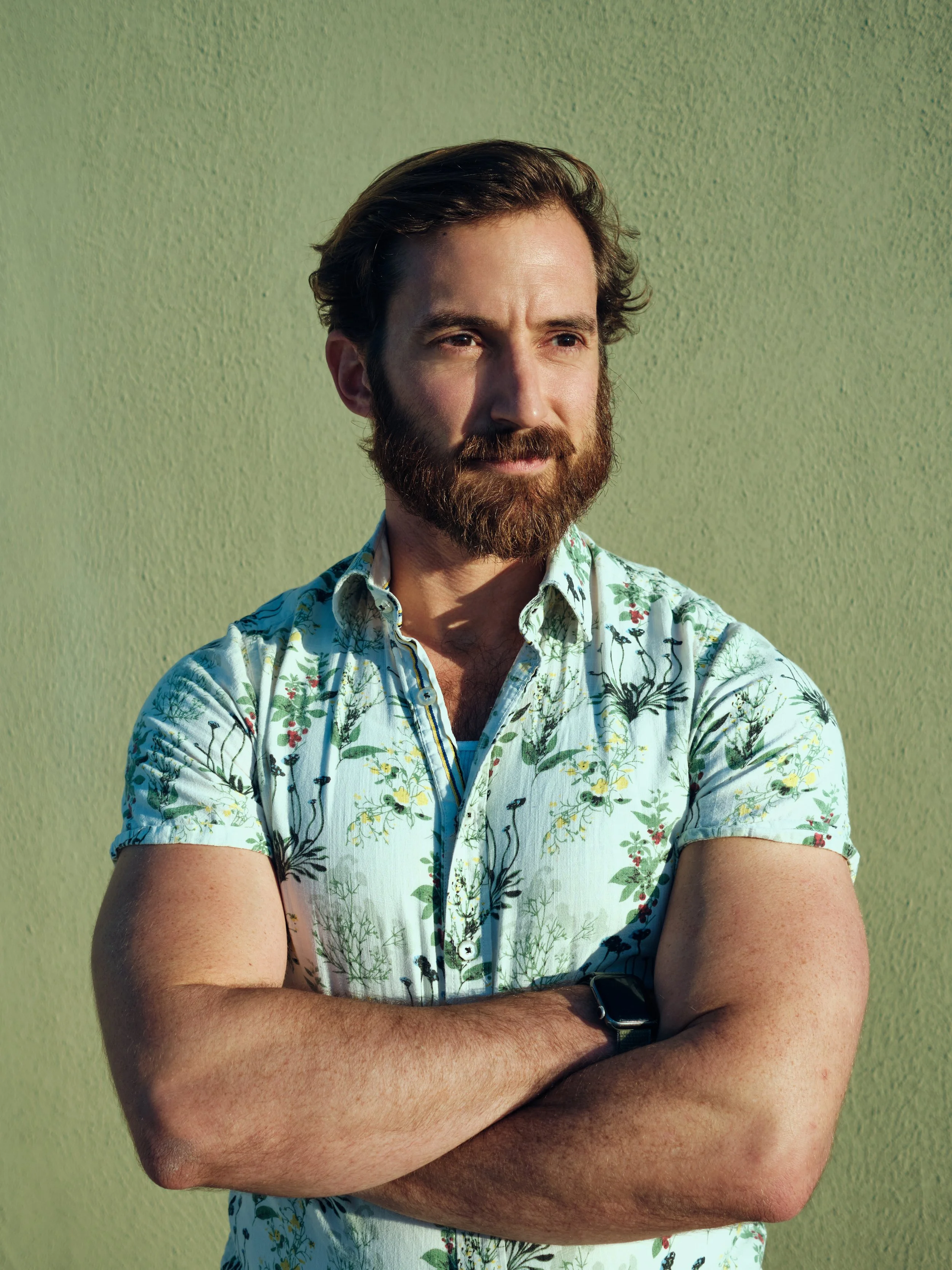 A man with a beard and brown hair standing against a light green wall, wearing a short-sleeved, floral button-up shirt and a black wristwatch, with arms crossed and looking off to the side.