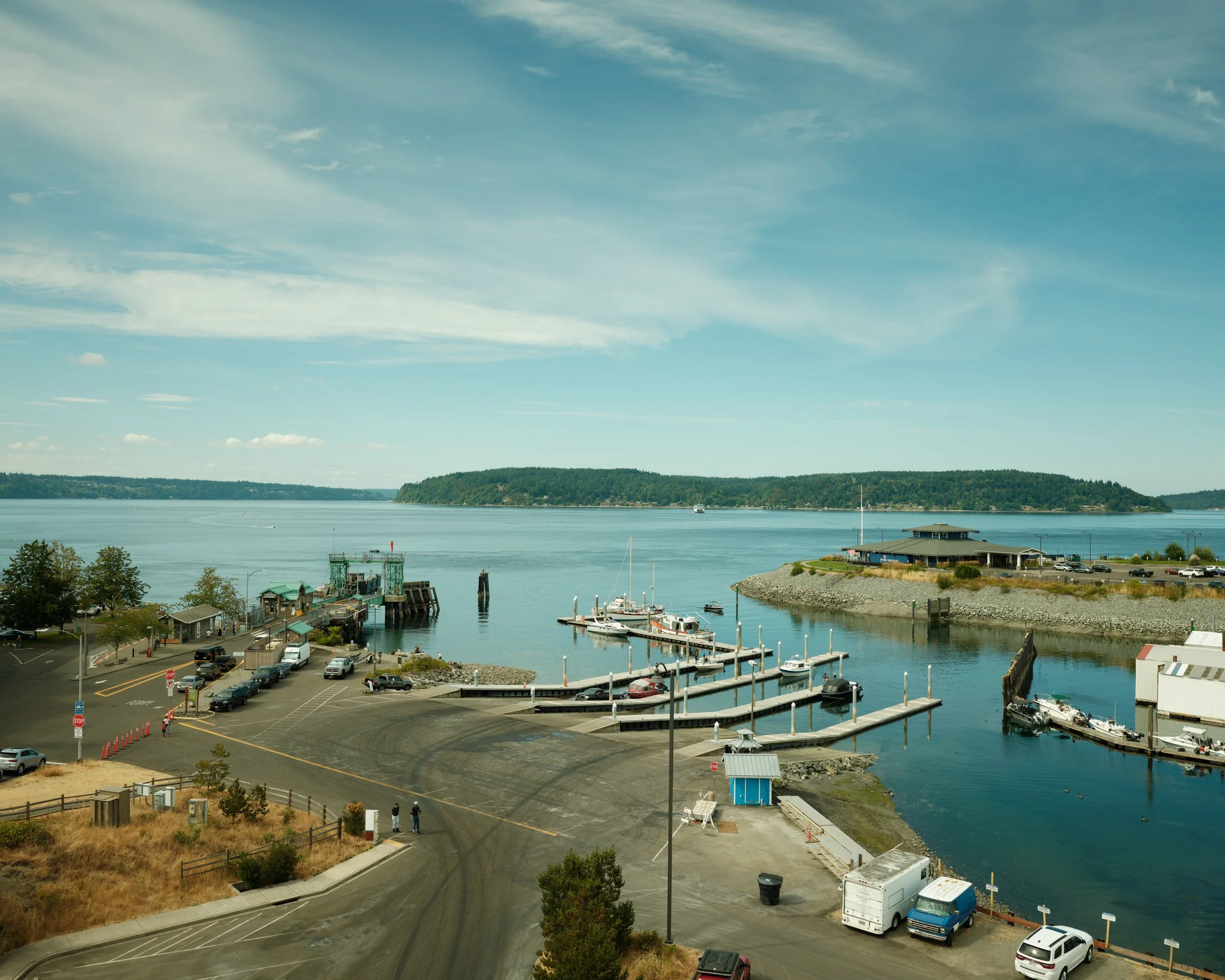 A harbor with boats docked at piers, a parking lot, and a large body of water with an island in the distance under a cloudy sky.