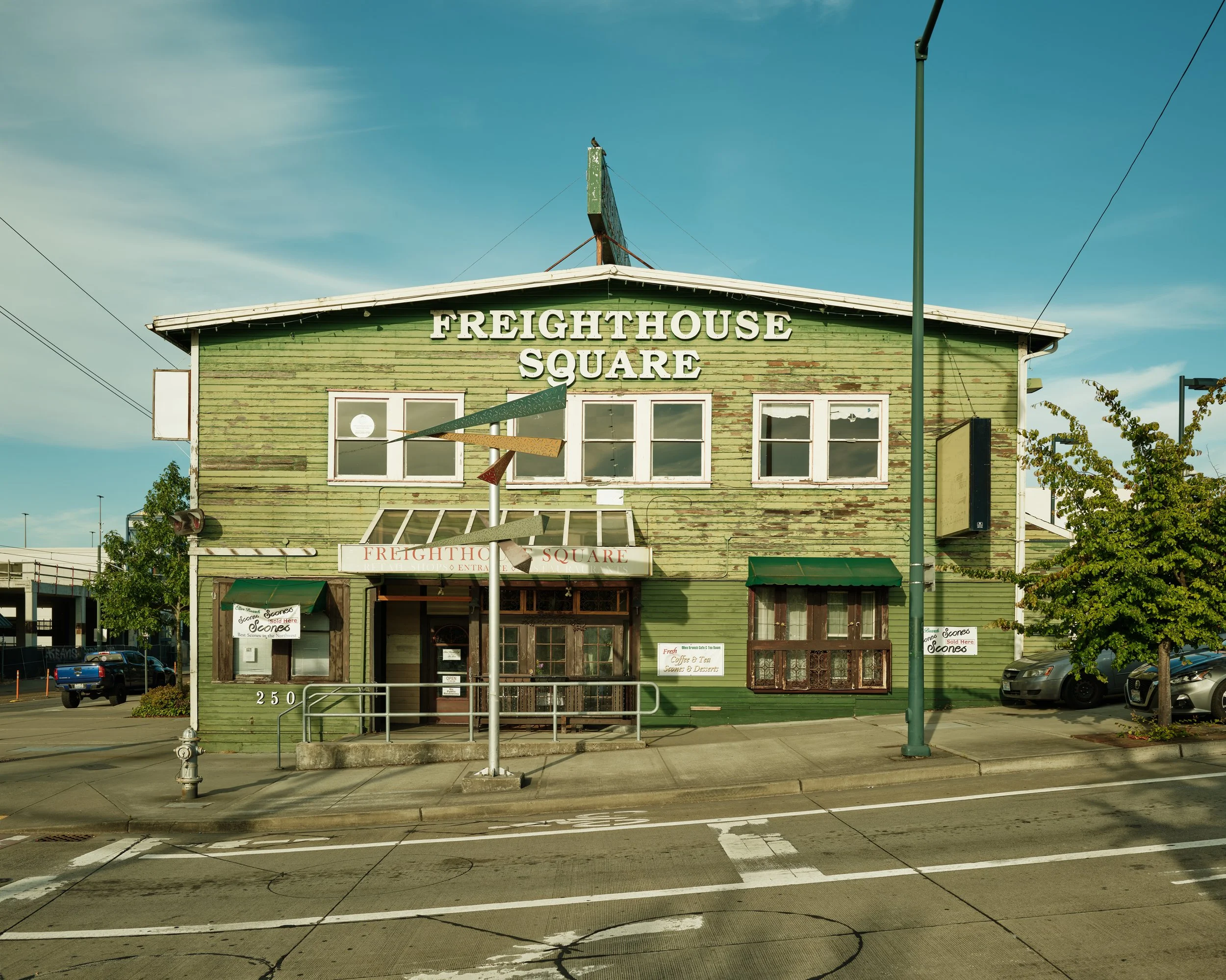 A green, weathered two-story building with the sign 'FREIGHTHOUSE SQUARE' on its front. The building has large windows, a small staircase, and multiple signs, including one that says 'Scones' and another advertising coffee and tea. There is a parking lot and street in front with a fire hydrant and a few parked cars, and a clear blue sky overhead.