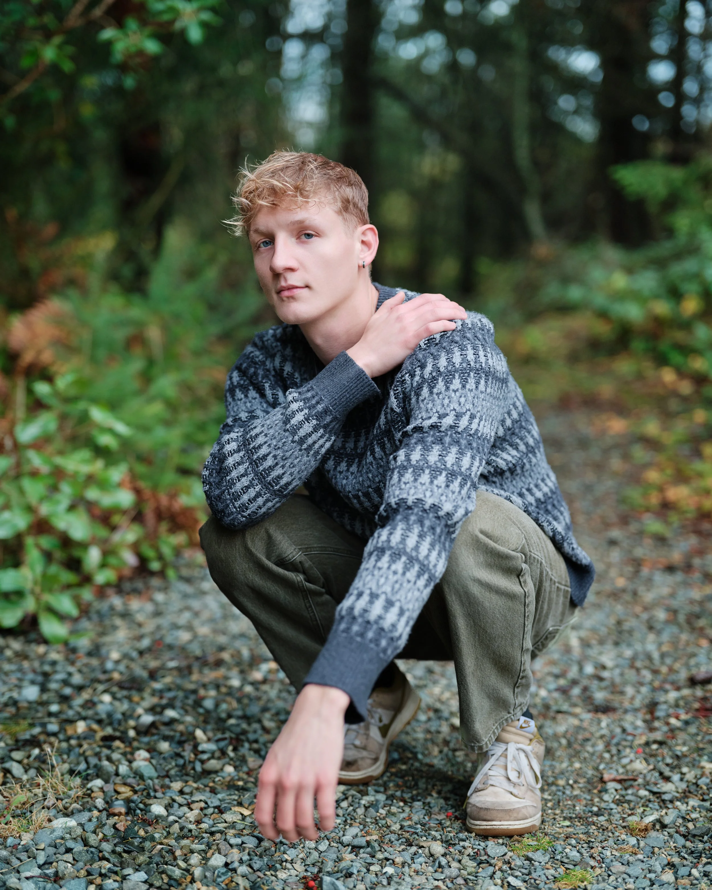 Young man squatting on a gravel path in a wooded area, wearing a patterned sweater, olive cargo pants, and beige sneakers.