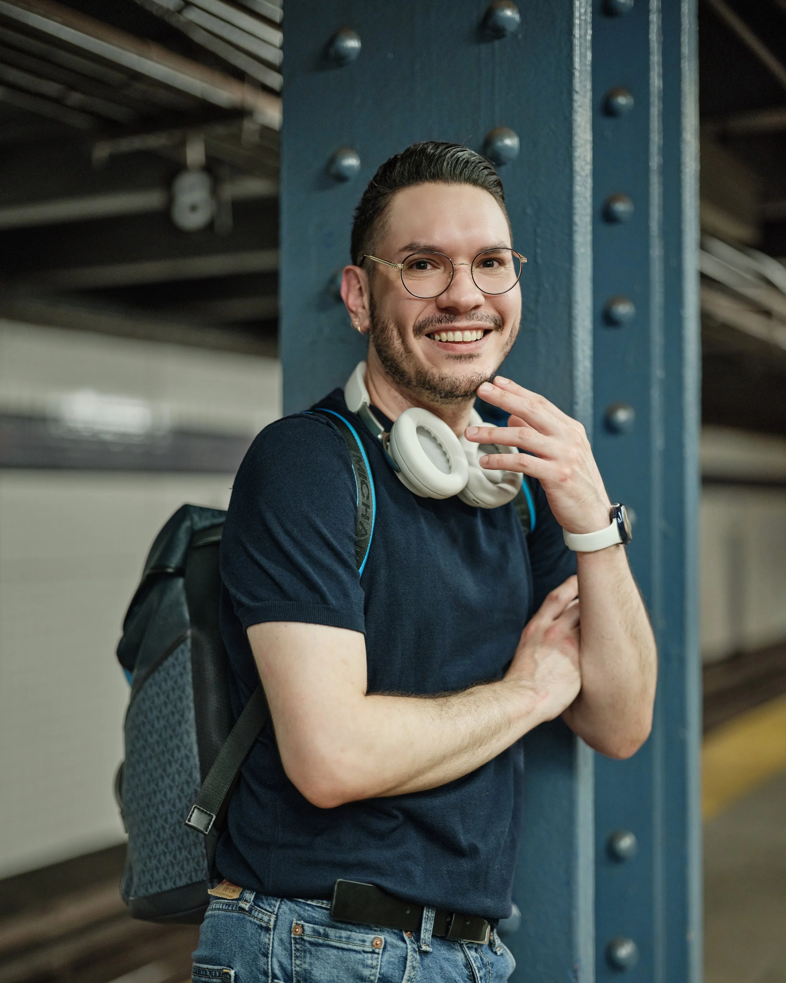 A young man with glasses and a beard smiling, standing at a subway station with a dark blue wall behind him. He's wearing a black t-shirt, carrying a backpack, and has a white headphone around his neck. He is posing with one hand near his chin and the other arm crossed over his chest.
