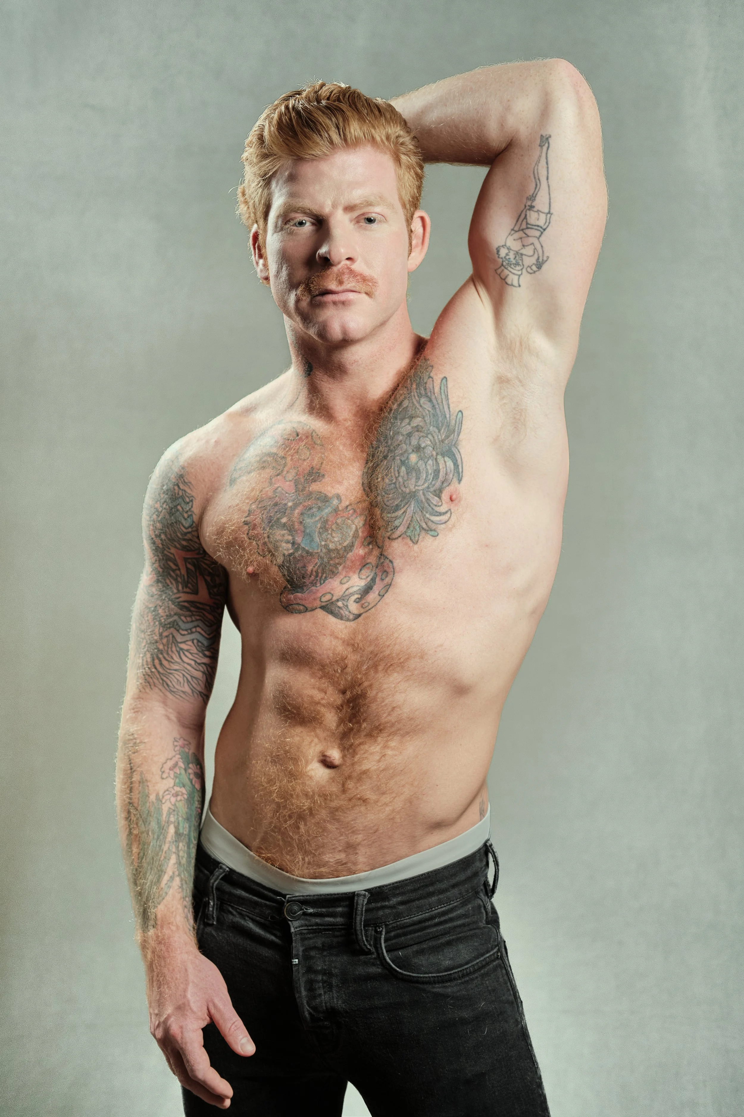 A shirtless man with red hair and tattoos posing with one arm behind his head, standing against a plain background.