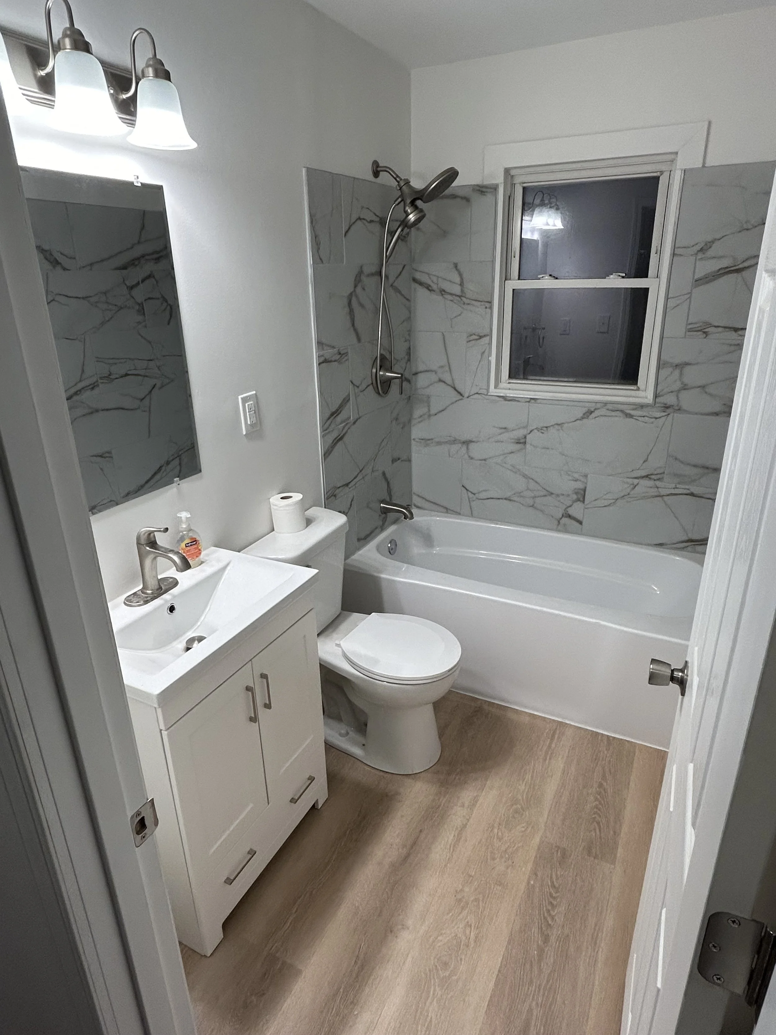 serving the Miami Valley, Dayton, and surrounding areas.  A small modern bathroom with a white vanity and sink, a toilet, a bathtub with a gray marble wall and a window, and a shower head above the bathtub.