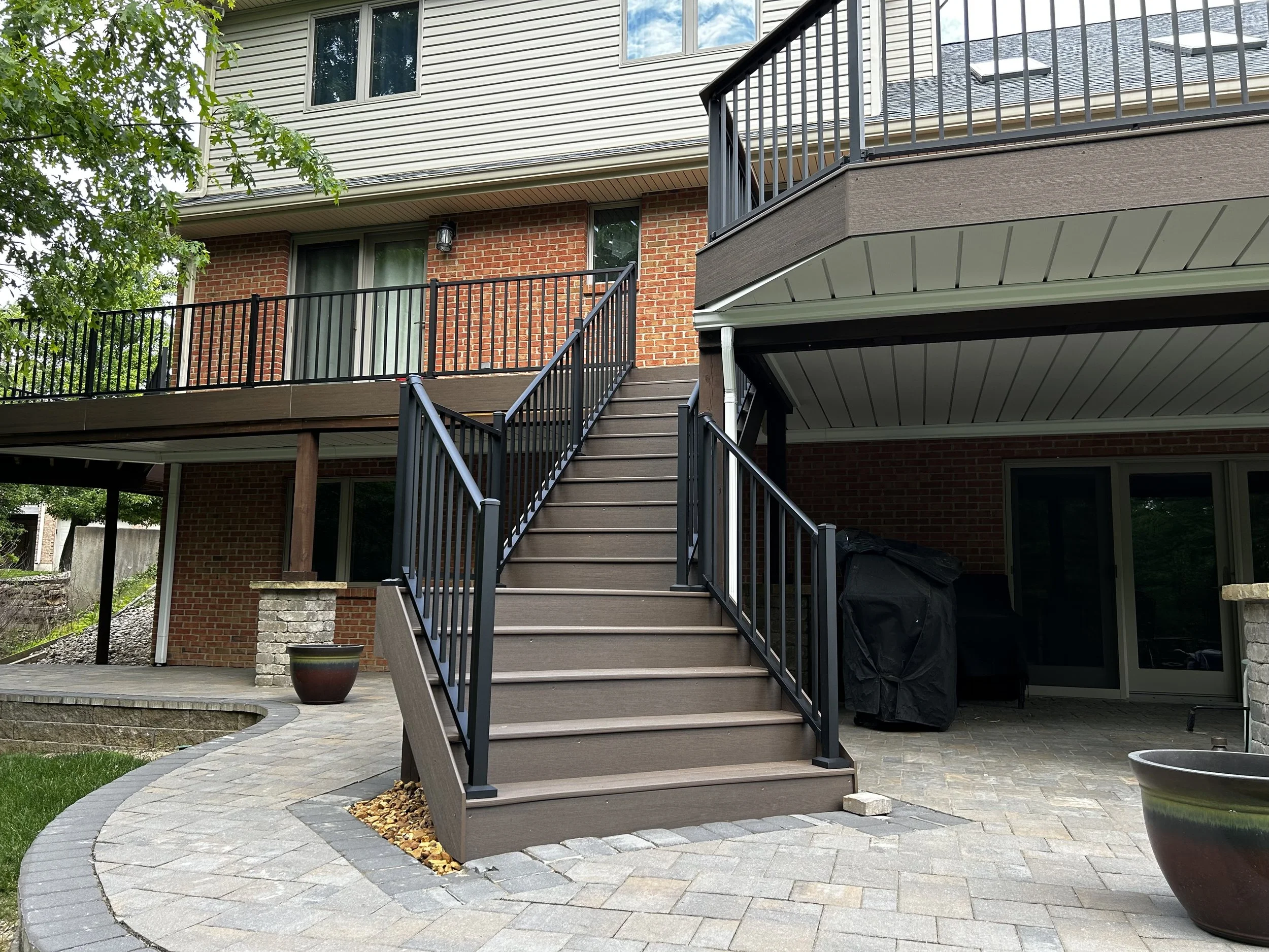 Outdoor view of a multi-level brick and siding house with a balcony, metal stairs, and a stone patio. There are potted plants and patio furniture under a covered area.