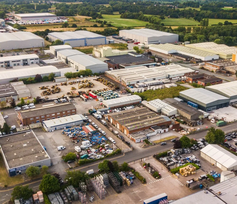 An aerial view of an industrial park with warehouses, trucks, construction equipment, and various vehicles surrounded by green fields.