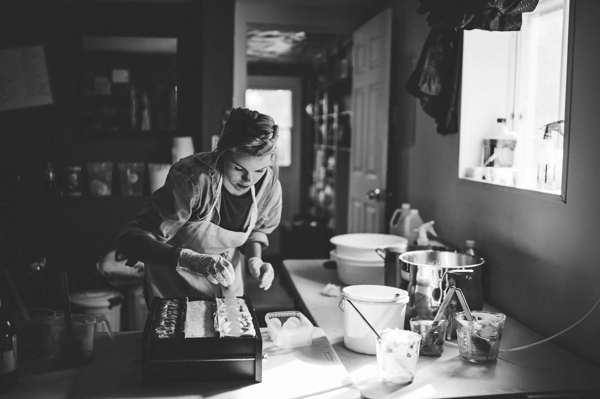 A woman in a kitchen, wearing gloves and an apron, is placing eggs into a baking pan, preparing baked goods on a countertop with various kitchen tools and ingredients visible.