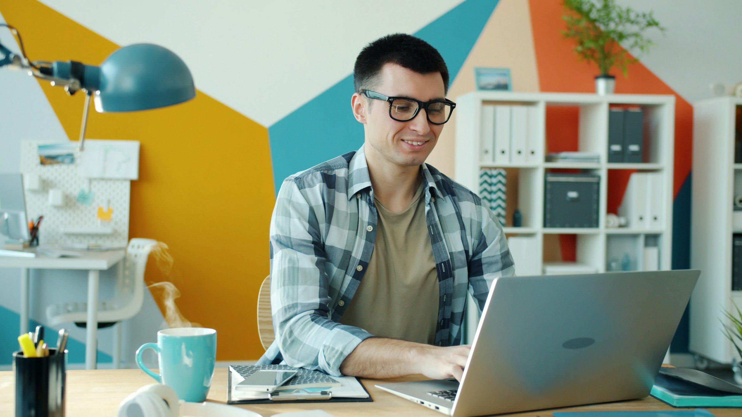 A young man with dark hair and glasses working on a laptop at a desk in a colorful office space.