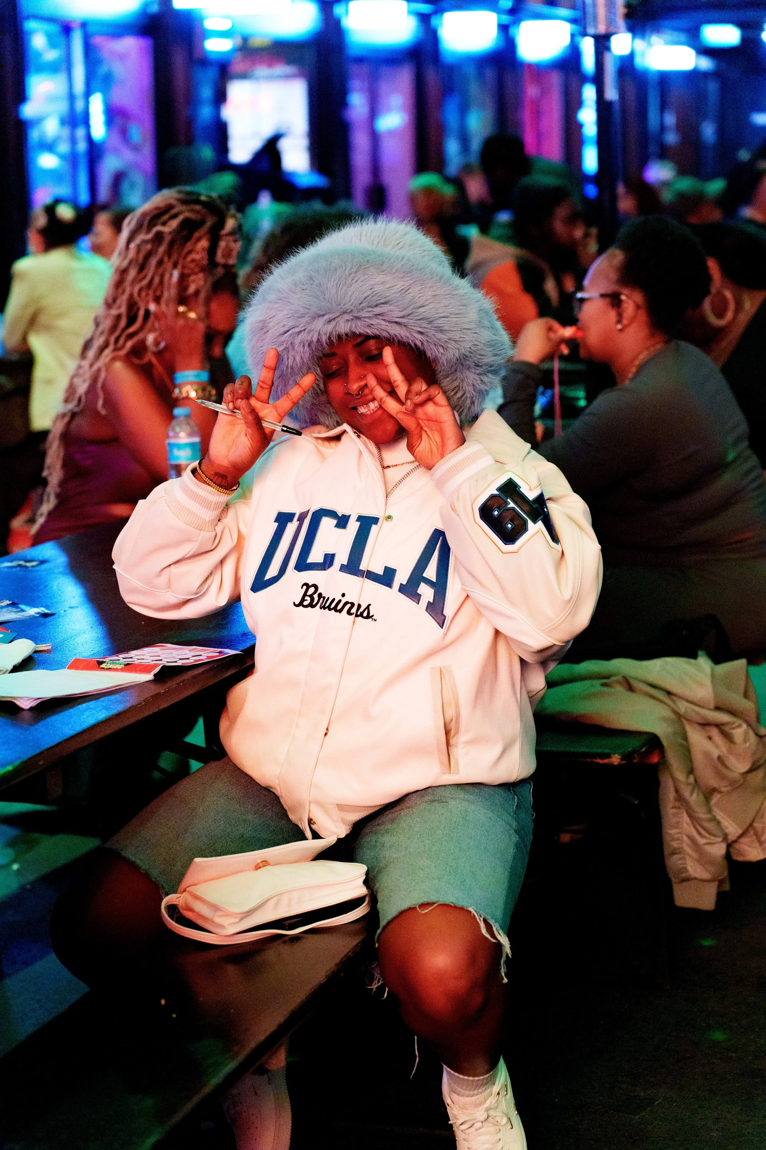 A young woman in a UCLA Bruins jacket with a fluffy blue hat makes a peace sign with both hands and smiles in a lively, crowded room with colorful lighting.