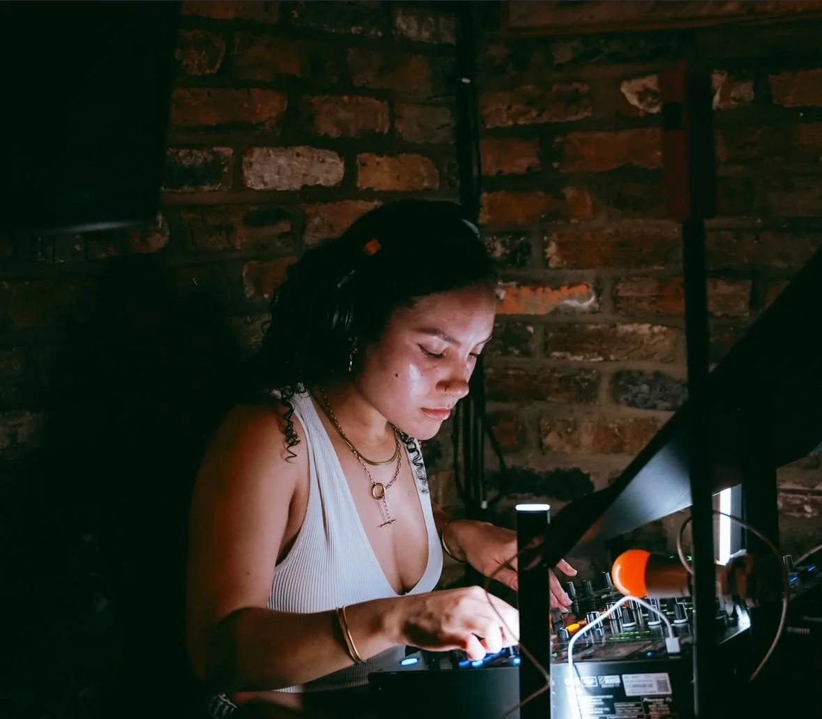 A woman DJ wearing headphones and a white tank top, focused on her equipment, with a brick wall in the background.